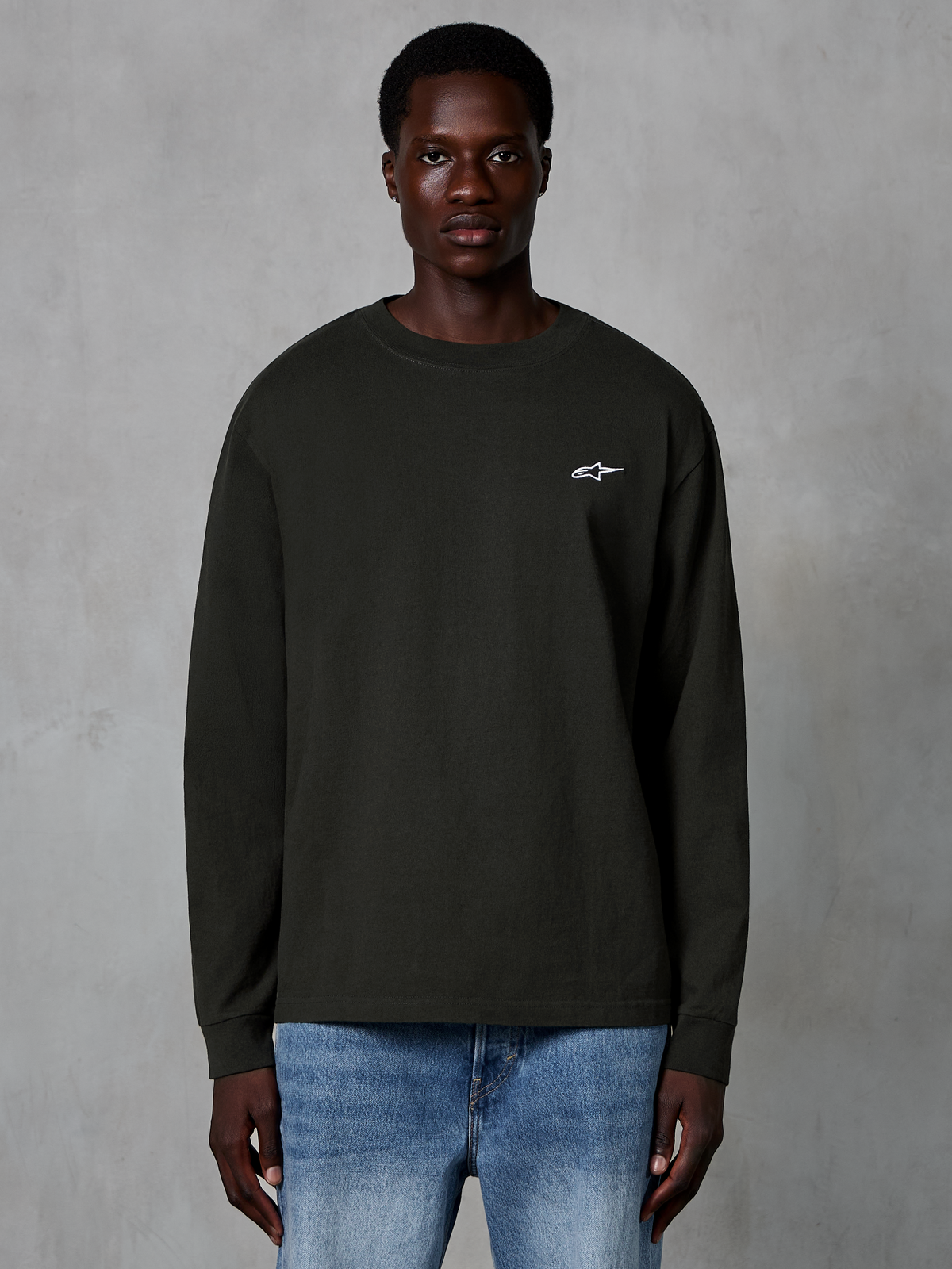 Alpinestars Essentials Ageless LS Tee, T-shirt met lange mouwen, Off Black, voorzien van een klein wit Alpinestars-logo op de borst, gedragen door een model met blauwe jeans, casual streetwear van biologisch katoen