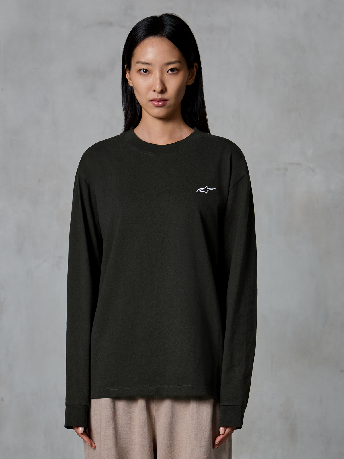 Model draagt Alpinestars Essentials Ageless LS Tee, T-shirt met lange mouwen, Off Black, met een klein wit Alpinestars-logo op de borst, gecombineerd met een beige broek, casual lifestyle streetwear