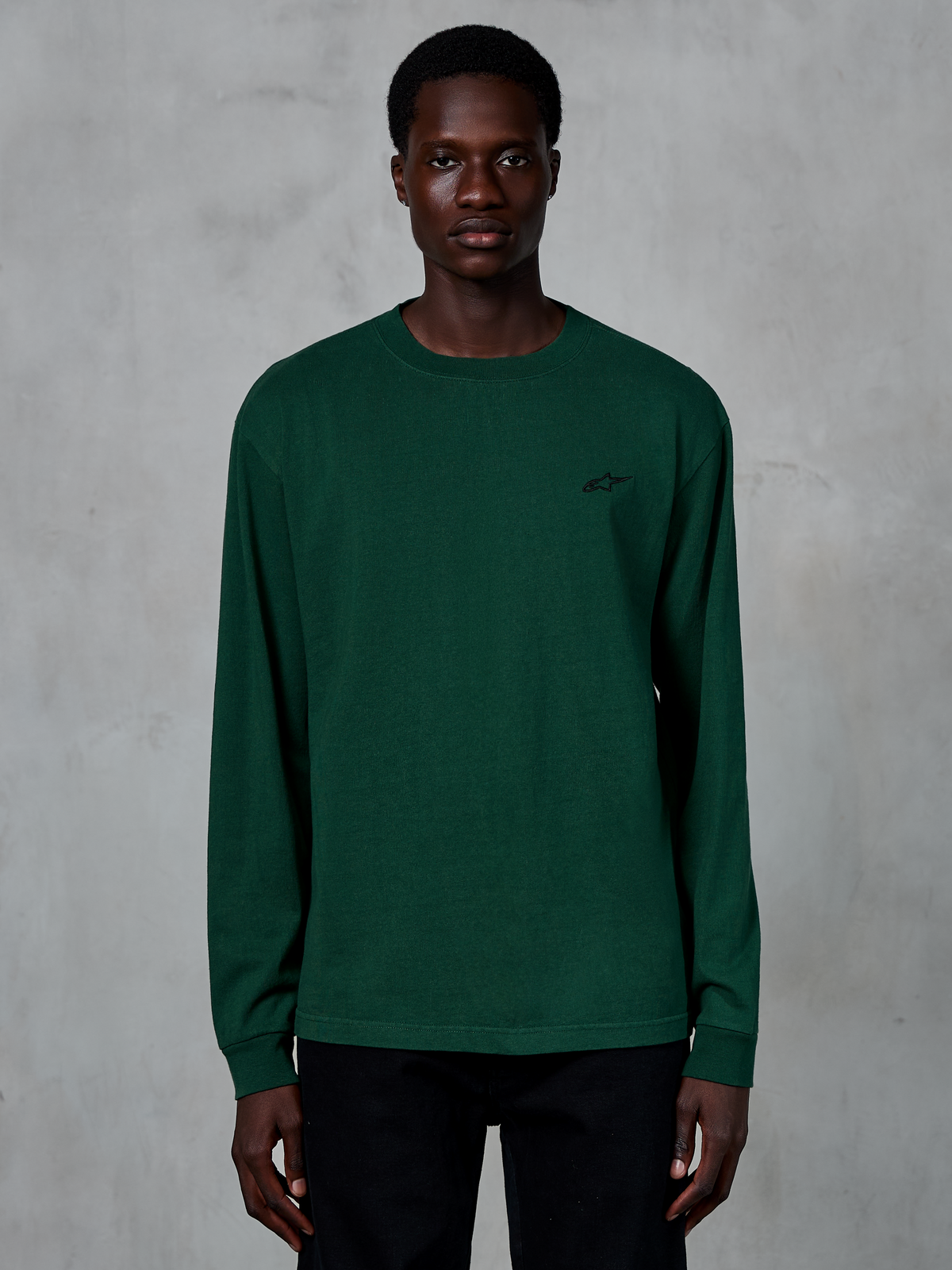 Alpinestars Essentials Ageless LS Tee, t-shirt à manches longues, vert sapin, porté par un mannequin, petit logo noir sur la poitrine, style streetwear décontracté, 100 % coton biologique avec finition délavée