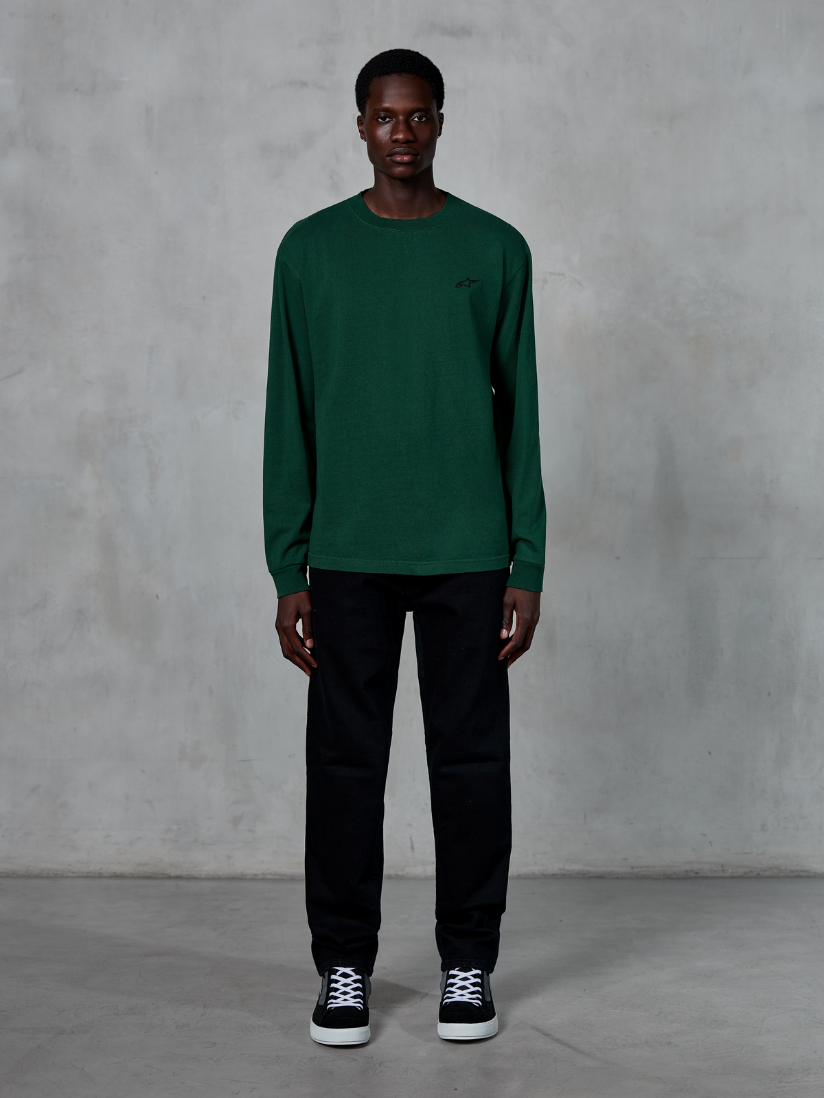 T-shirt à manches longues Alpinestars Essentials Ageless, vert sapin, doté d'un petit logo noir sur la poitrine, porté par un mannequin avec un pantalon noir et des baskets, style streetwear décontracté
