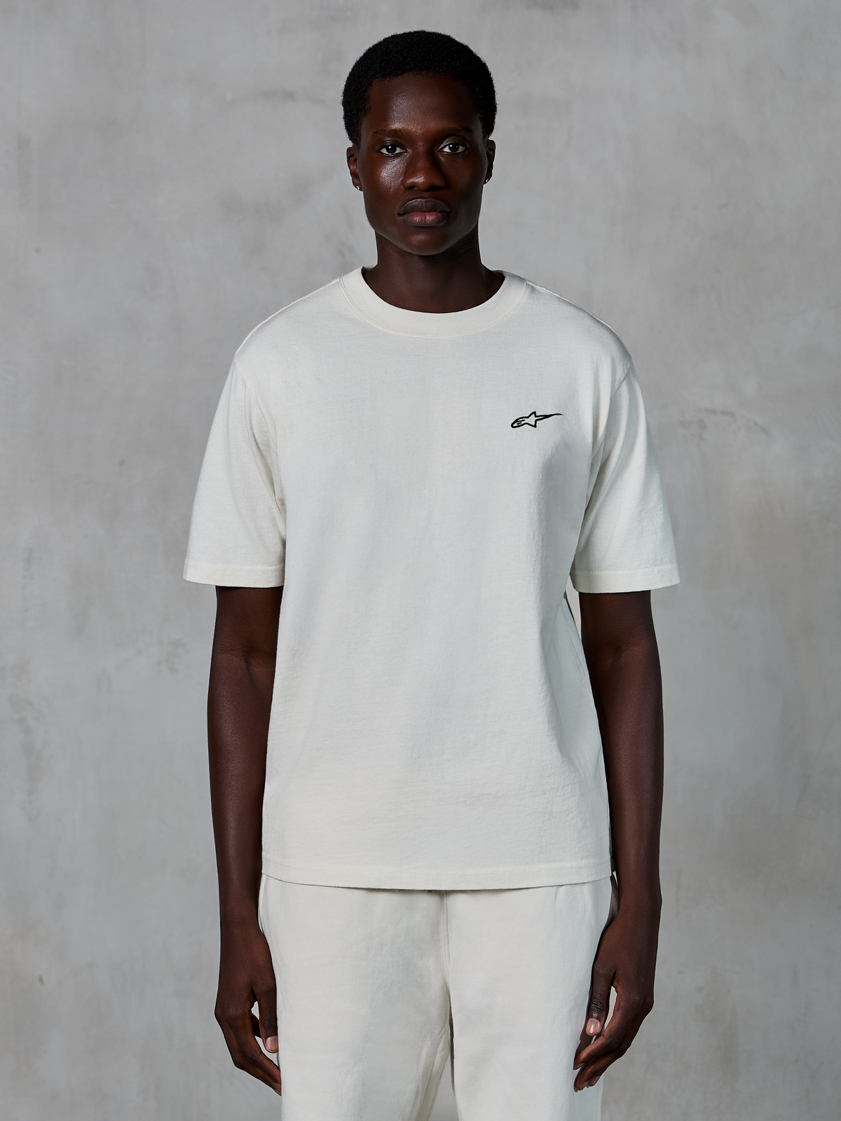 Alpinestars Essentials Ageless SS Tee, t-shirt à manches courtes, blanc cassé, porté par un mannequin homme, doté d'un petit logo Alpinestars noir sur la poitrine, coupe décontractée, jersey de coton biologique, style streetwear motorsport décontracté