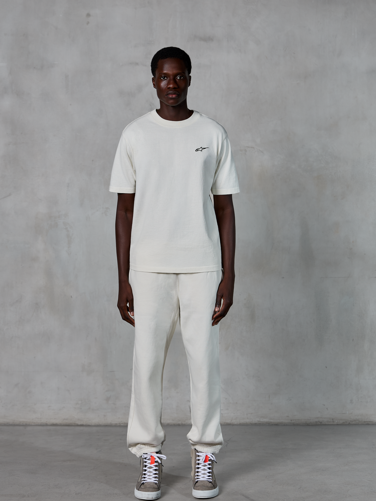 T-shirt Alpinestars Essentials Ageless SS, t-shirt à manches courtes, blanc cassé, arborant un petit logo Alpinestars noir sur la poitrine, porté avec un pantalon de jogging et des baskets assortis pour un look décontracté