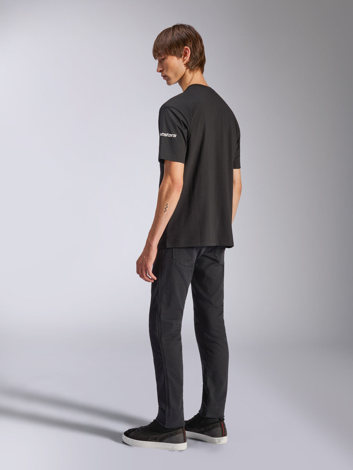 Alpinestars Frontal CSF T-Shirt, kurzärmeliges T-Shirt, Schwarz, weißes Astars-Branding auf dem Ärmel, getragen von einem Model mit schwarzer Hose und Schuhen, lässiges, vom Motorsport inspiriertes Design