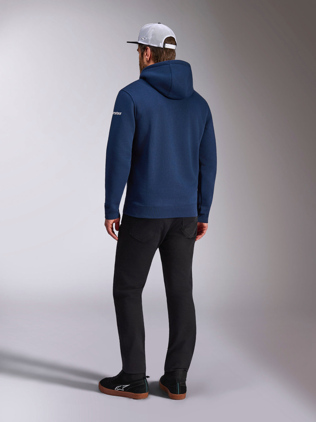 Sweat à capuche Alpinestars Frontal, vue arrière d'un sweat à capuche en polaire bleu marine, logo en lettres blanc sur la manche, porté avec un pantalon noir et une casquette trucker blanche en maille filet, streetwear décontracté inspiré du sport automobile
