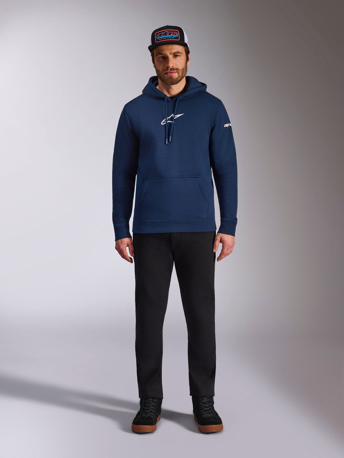 Modèle masculin portant le sweat à capuche Frontal Alpinestars bleu marine, polaire à enfiler avec logo Astars blanc sur la poitrine et logo lettrage sur la manche, doté d'une poche kangourou et d'une capuche à cordon, associé à un pantalon noir et une casquette de marque, streetwear motorsport décontracté