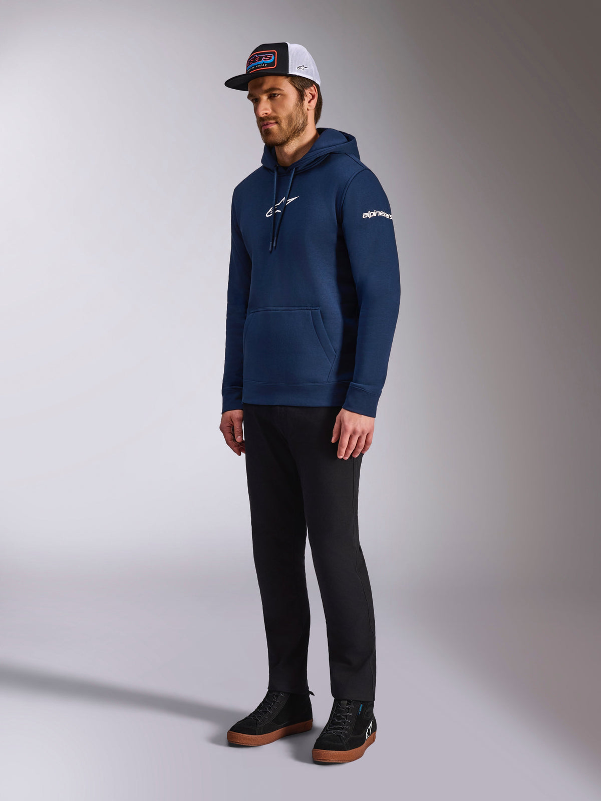 Sweat à capuche Frontal Alpinestars en bleu marine, sweat en polaire décontracté avec logo Astars blanc sur la poitrine et logo en lettres sur la manche, porté avec une casquette trucker et un pantalon noir, vêtement lifestyle moderne inspiré du sport automobile