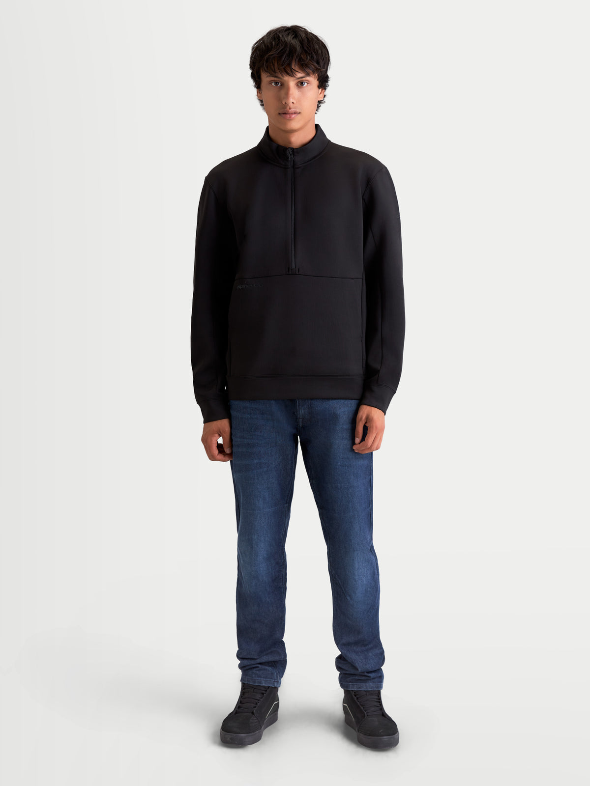 Alpinestars Sweat Half-Zip Apera, pull sportswear pour homme, noir, avec fermeture demi-zippée et poche kangourou, style inspiré du sport automobile pour un confort décontracté, porté par un modèle avec un jean bleu.