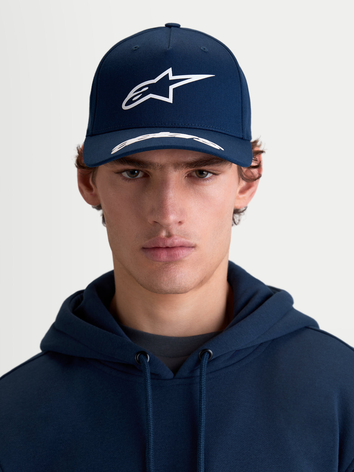Alpinestars Arclight pet, sportswear-pet met gebogen klep, donkerblauw en wit, vooraanzicht op model, voorzien van een gestructureerde kroon, getextureerd puffprint Astars-logo en premium katoenen keperstof.