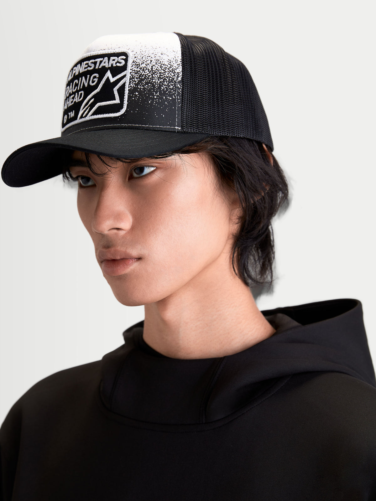 Alpinestars Gravel Trucker pet, casual sportswear pet op model, zwart en wit, voorzien van een gestructureerde kroon, gebogen klep, ademende mesh achterzijde en geweven patch met grindtextuur en Alpinestars Racing Ahead-branding.