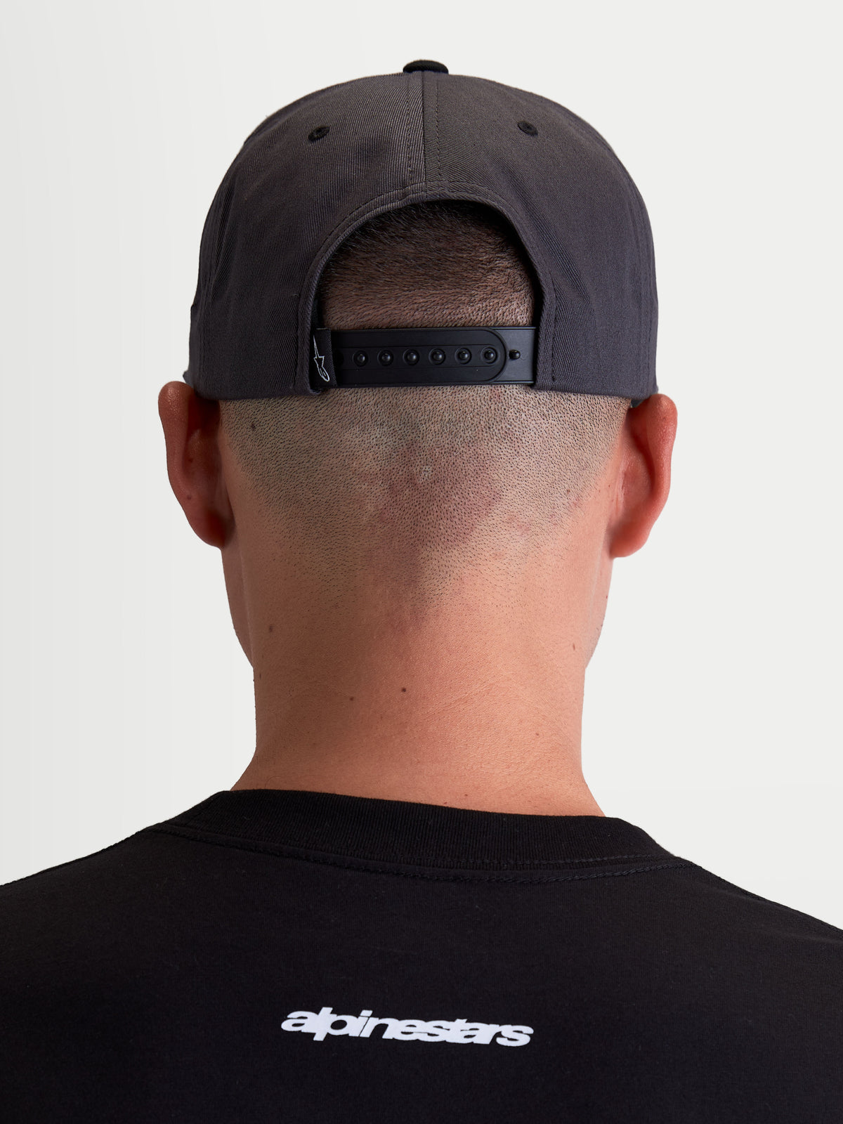 Alpinestars Stadium Snapback pet, achteraanzicht op model met de verstelbare snapback-sluiting en gestructureerde kroon, donkergrijs en zwart, duurzame keperstof, casual sportswear.