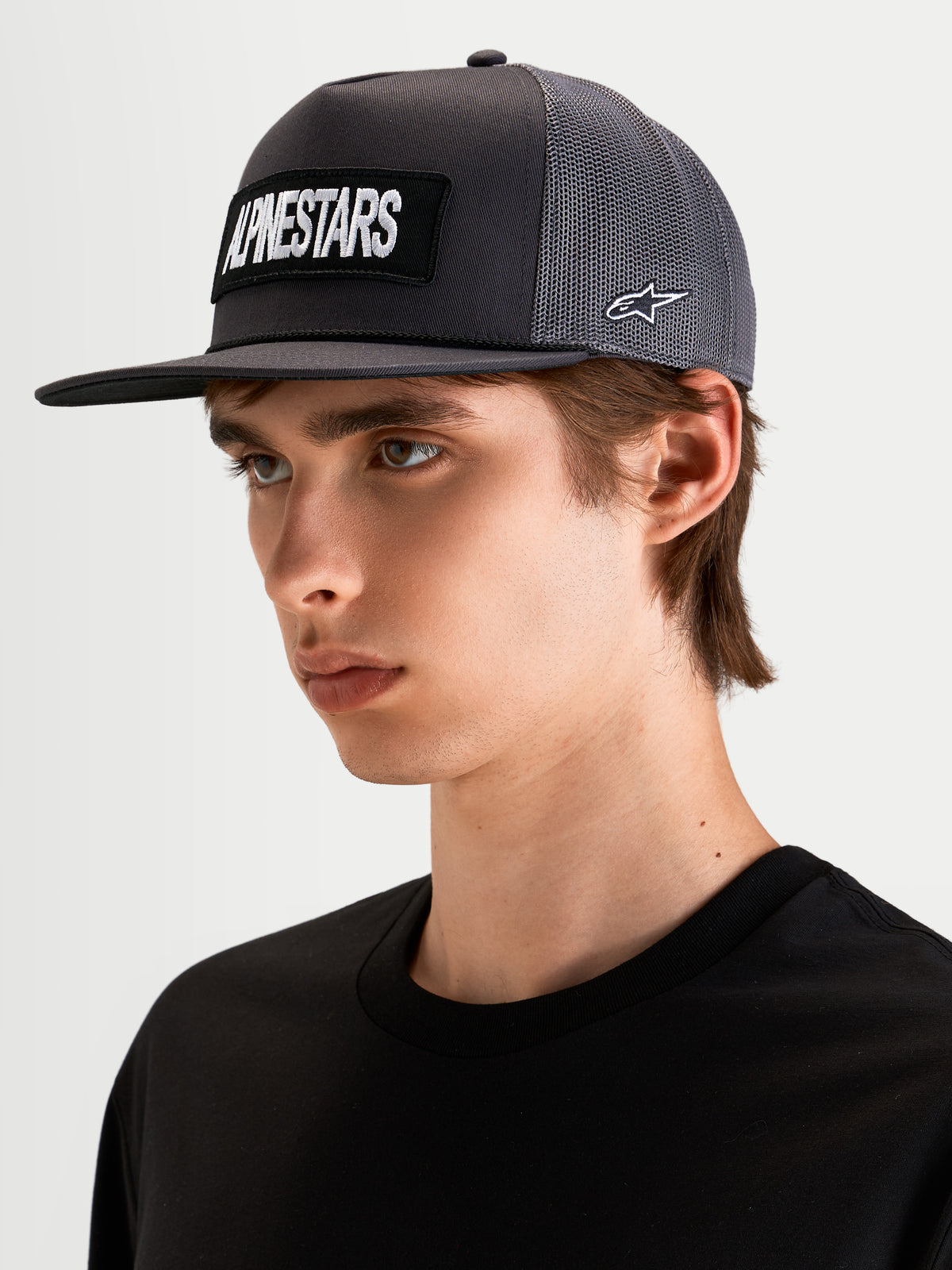 Alpinestars Casquette Trucker Ten, casquette sportswear à visière plate en gris foncé, portée par un modèle, avec un écusson brodé du logo Alpinestars à l'avant et une broderie du logo Astars sur le côté