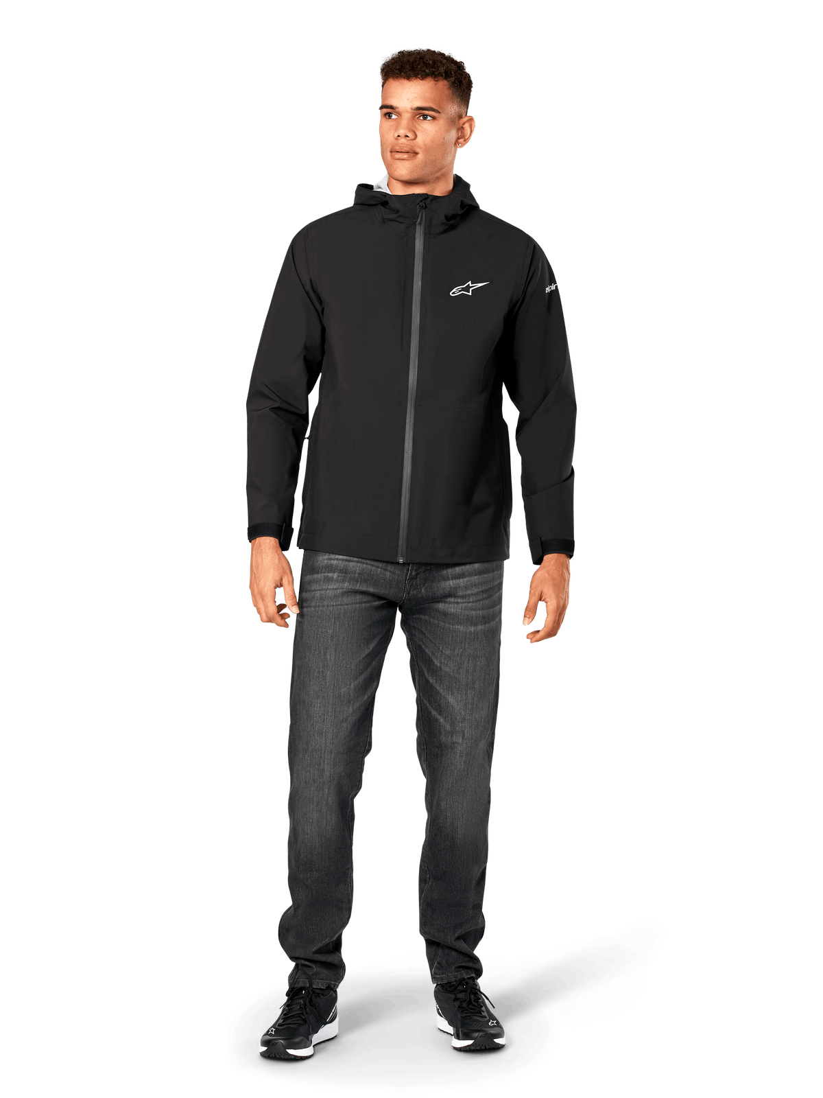 Alpinestars Kascade Regenjacke, Herren-Regenjacke, Schwarz, kombiniert mit dunkelgrauen Jeans und schwarzen Sneakern, mit Kapuzendesign, beschichtetem Frontreißverschluss und weißem Alpinestars-Logo auf der Brust, lässige, vom Motorsport inspirierte Streetwear