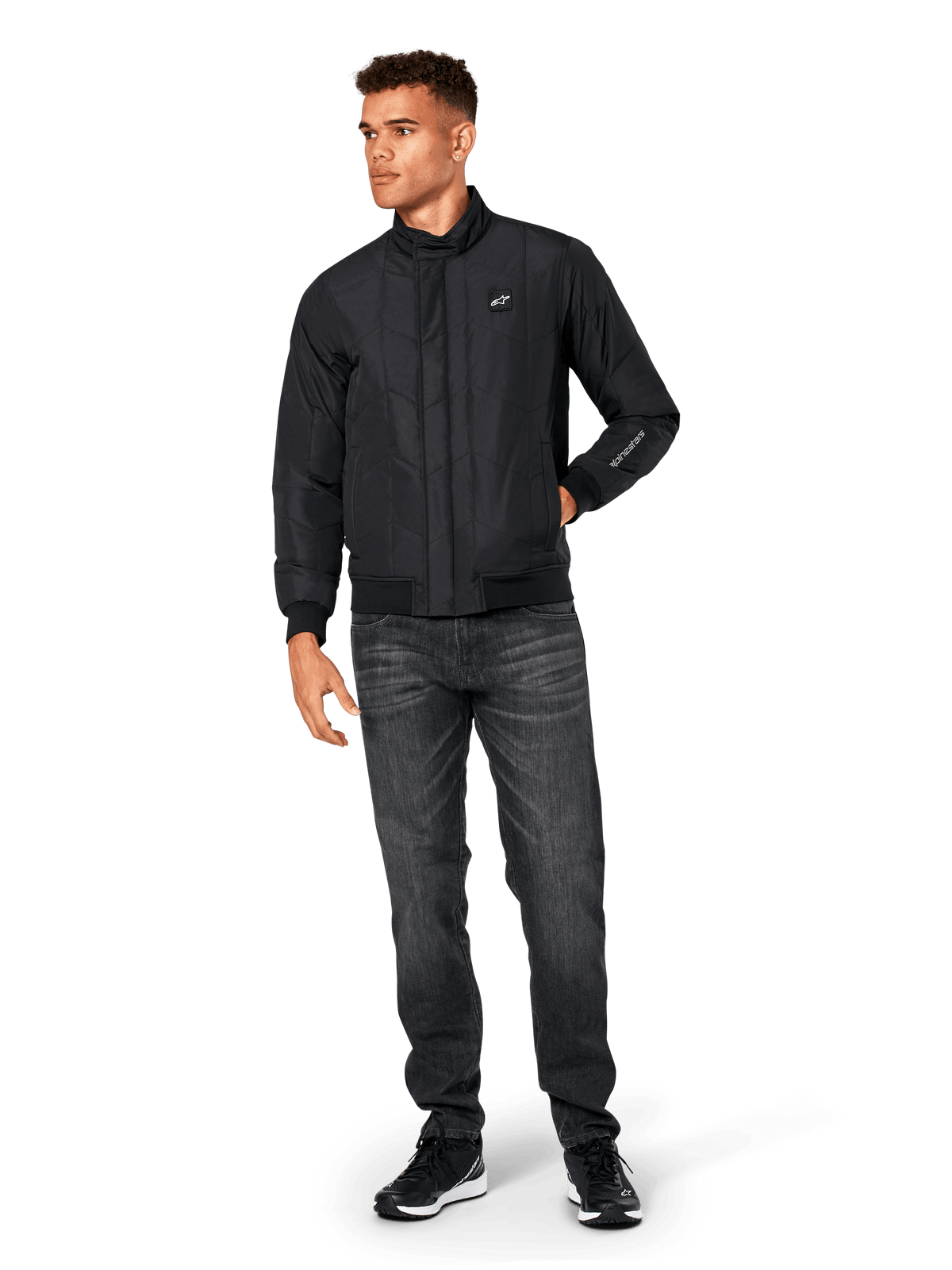 Alpinestars Precedent Jacke, gesteppte Sportjacke, Schwarz, getragen mit dunkelgrauer Jeans und schwarzen Sneakern, mit schrägem Steppmuster, niedrigem Stehkragen, Rippstrickbündchen an Ärmeln und Saum, weißem Logo-Patch auf der Brust und Stickerei am Ärmel.