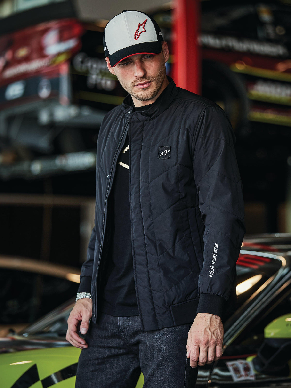 Alpinestars Precedent Jacke, schwarze Steppjacke im Formel-1-Stil, mit einem ikonischen schrägen Steppmuster, Logo-Patch auf der Brust und weißer Ärmelstickerei, getragen mit einer passenden Alpinestars-Kappe in einer Motorsport-Garage.