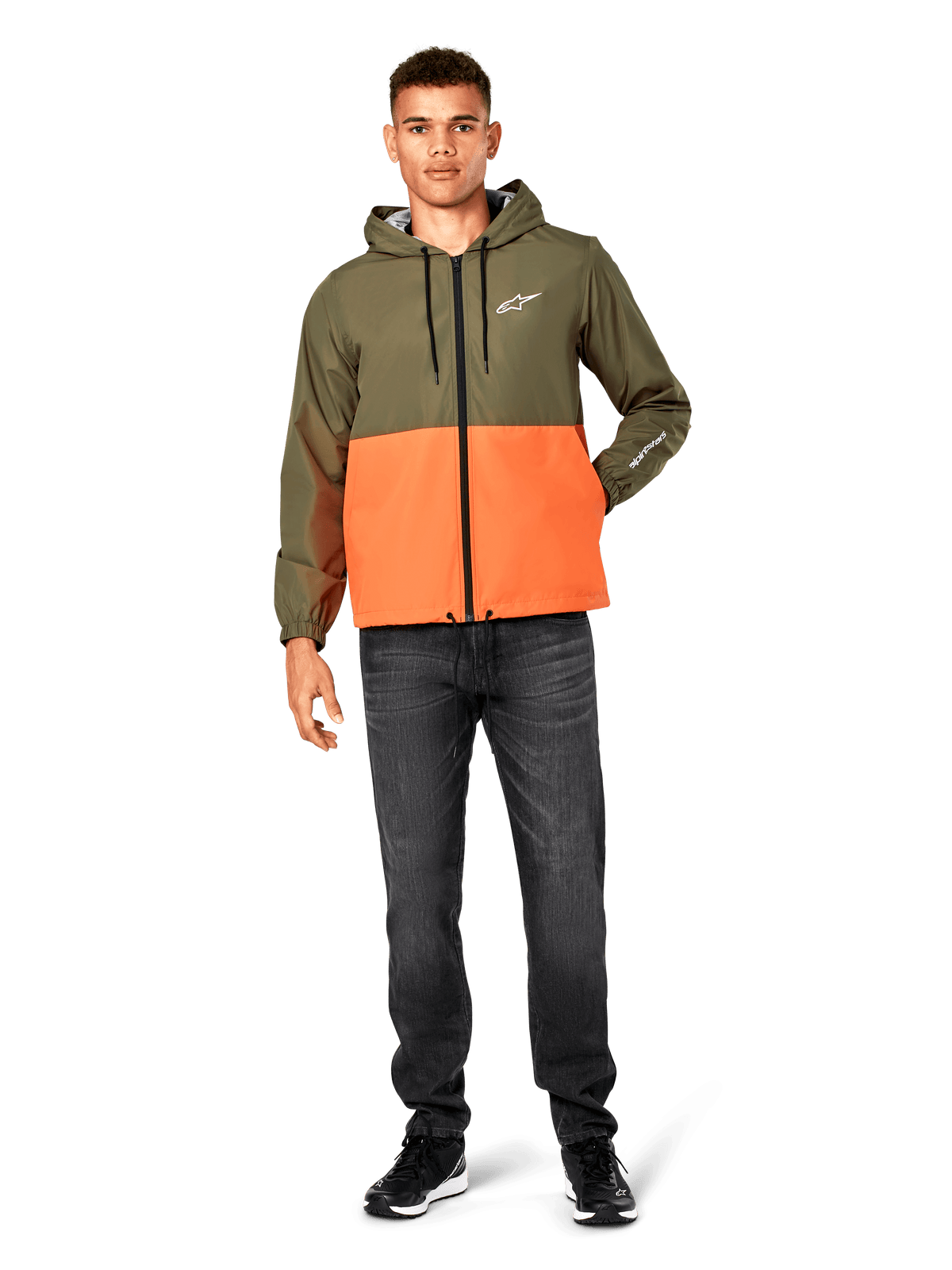 Veste coupe-vent Alpinestars Speeder, bicolore vert militaire et orange, veste à capuche légère avec cordons de serrage noirs et fermeture éclair avant, logo blanc sur la poitrine et la manche, portée avec un jean foncé et des baskets noires, style sportswear décontracté