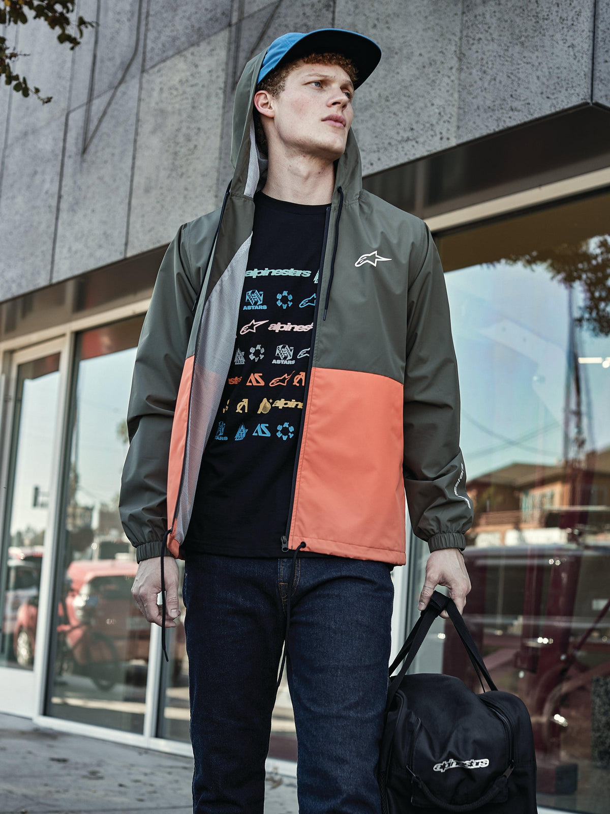 Man in Alpinestars Speeder Windbreaker Jas in Military Orange, kleurgeblokt ontwerp in legergroen en oranje, capuchon met meshvoering, gecombineerd met zwart T-shirt met grafisch logo, blauwe jeans en een zwarte Alpinestars duffelbag, casual sportswear lifestyle-look