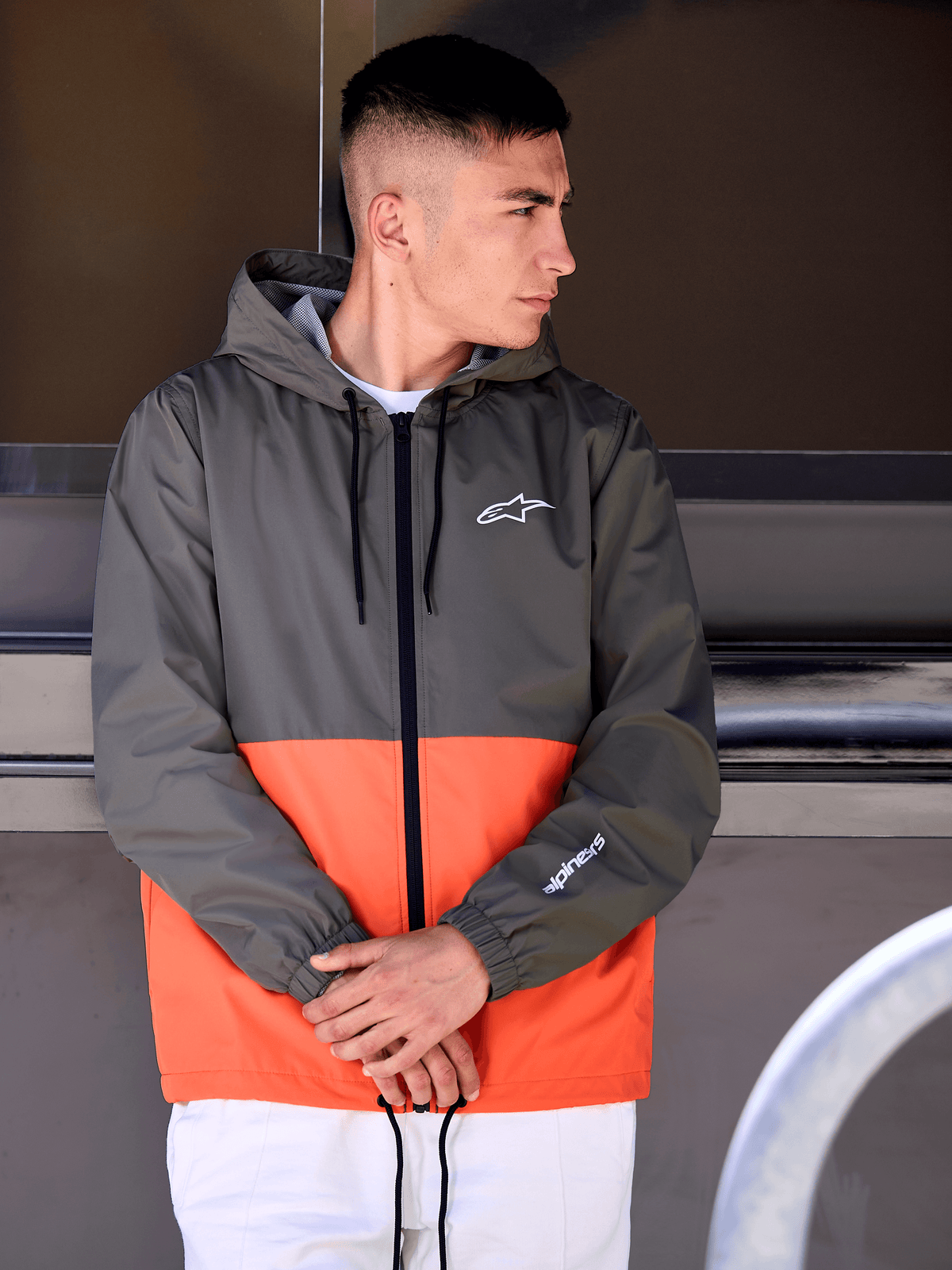 Homme portant la veste coupe-vent Alpinestars Speeder, design color-block orange militaire et vert foncé, vêtement d'extérieur décontracté à capuche avec logos blancs sur la poitrine et la manche, associé à un pantalon blanc pour un look lifestyle inspiré du sport automobile