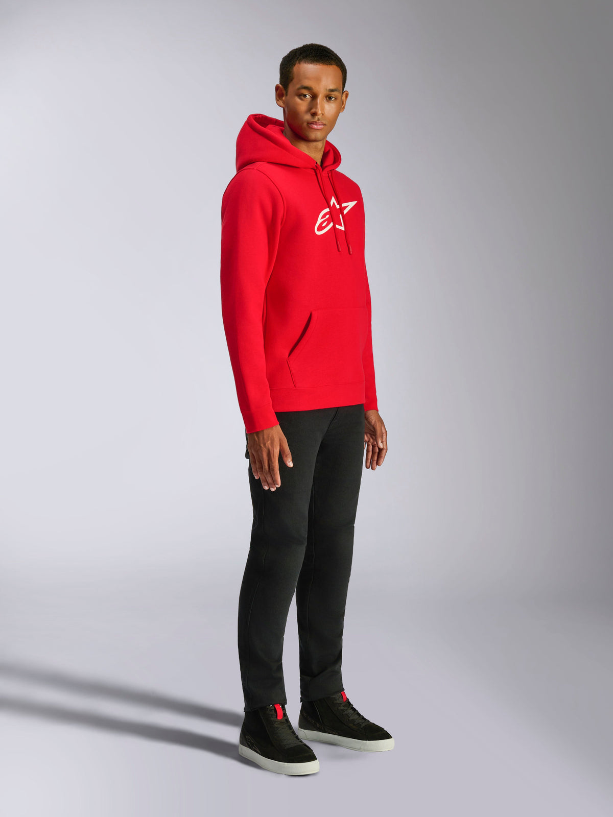 Alpinestars Ageless V3 Hoodie, rood met wit geborduurd logo, casual sportieve hoodie gedragen door een man met zwarte broek en zwarte sneakers, voorzien van een capuchon met trekkoord en geribde boorden