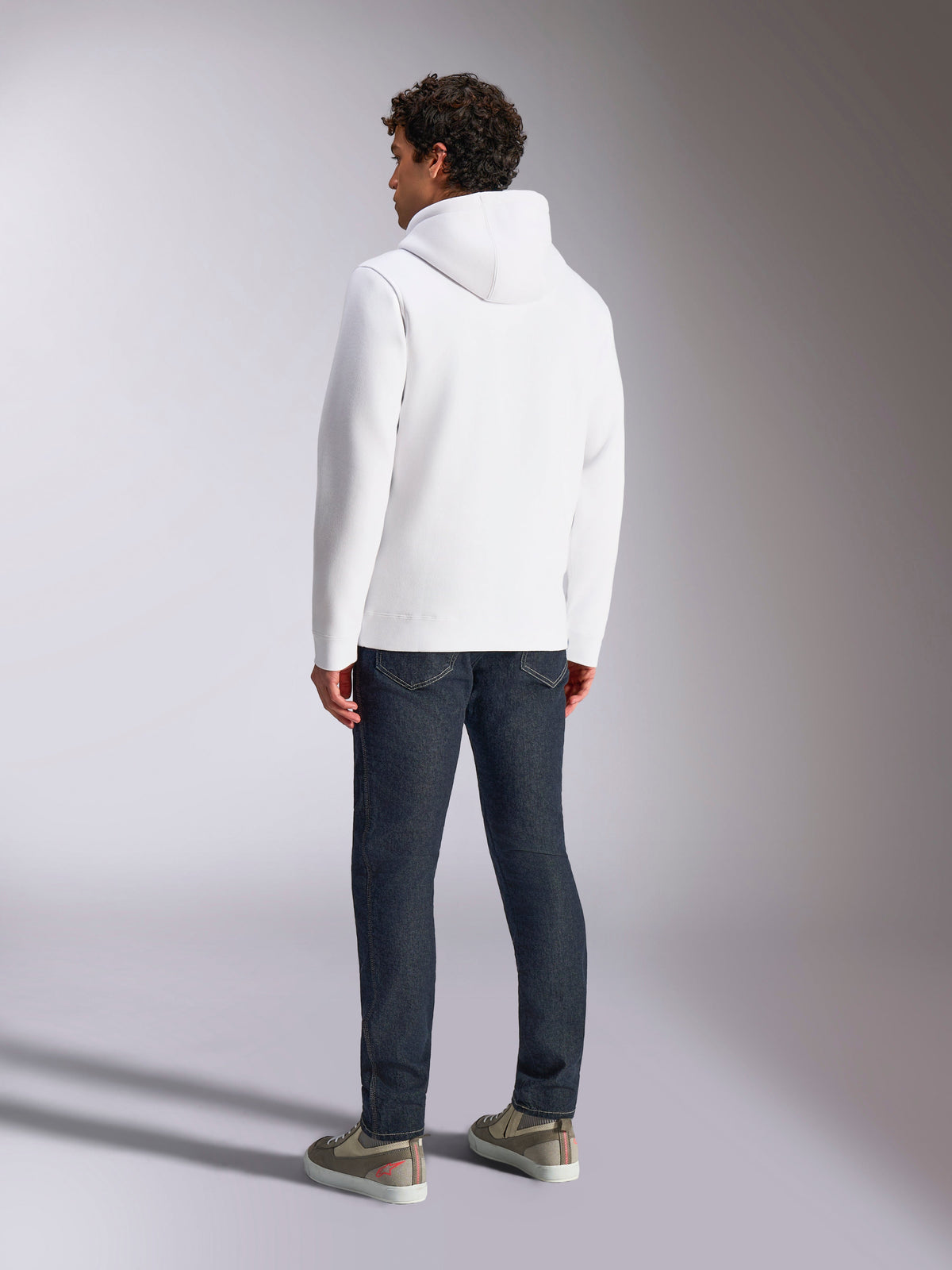 Alpinestars Blaze V3 Hoodie, vue arrière d'un mannequin portant un sweat à capuche blanc, style sportswear décontracté avec un jean bleu foncé et des baskets grises, design moderne inspiré des sports mécaniques
