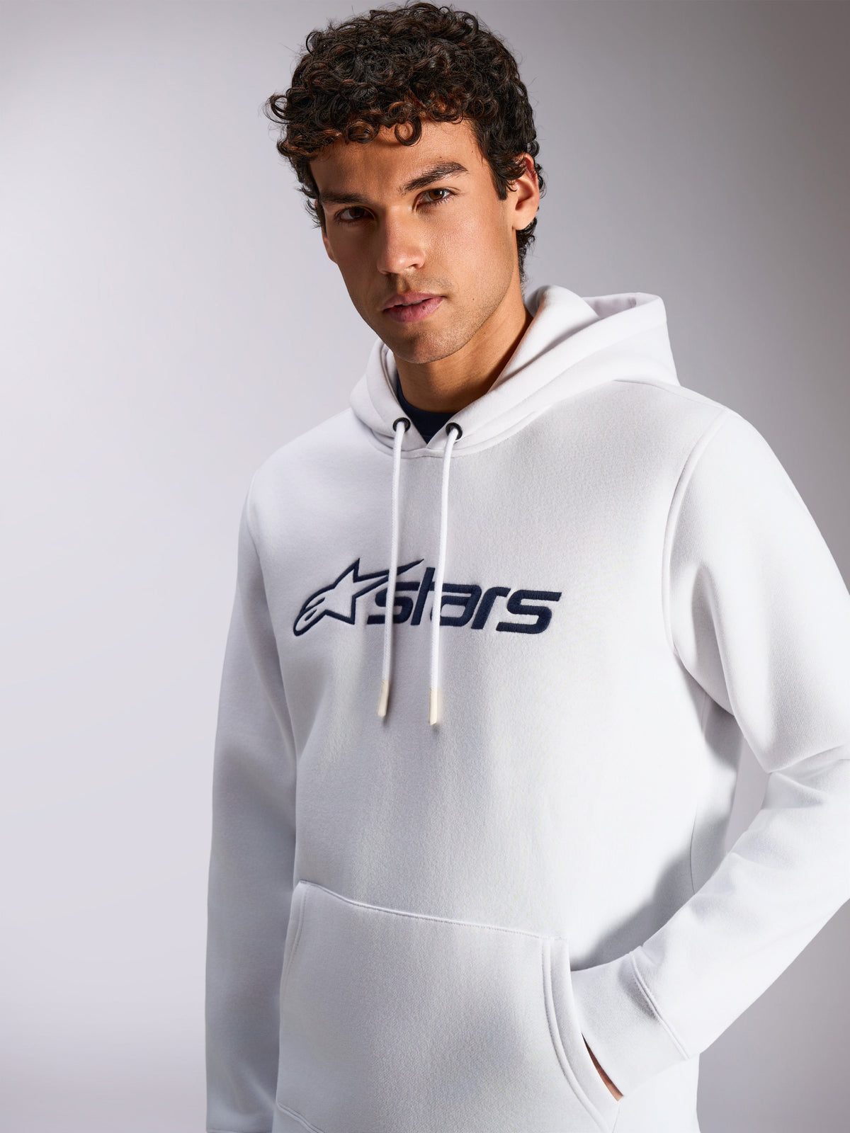 Sweat à capuche Alpinestars Blaze V3, blanc et bleu marine, porté par un mannequin homme, sportswear décontracté, logo Alpinestars bleu marine sur le devant, capuche avec cordon de serrage, poche kangourou, design lifestyle moderne