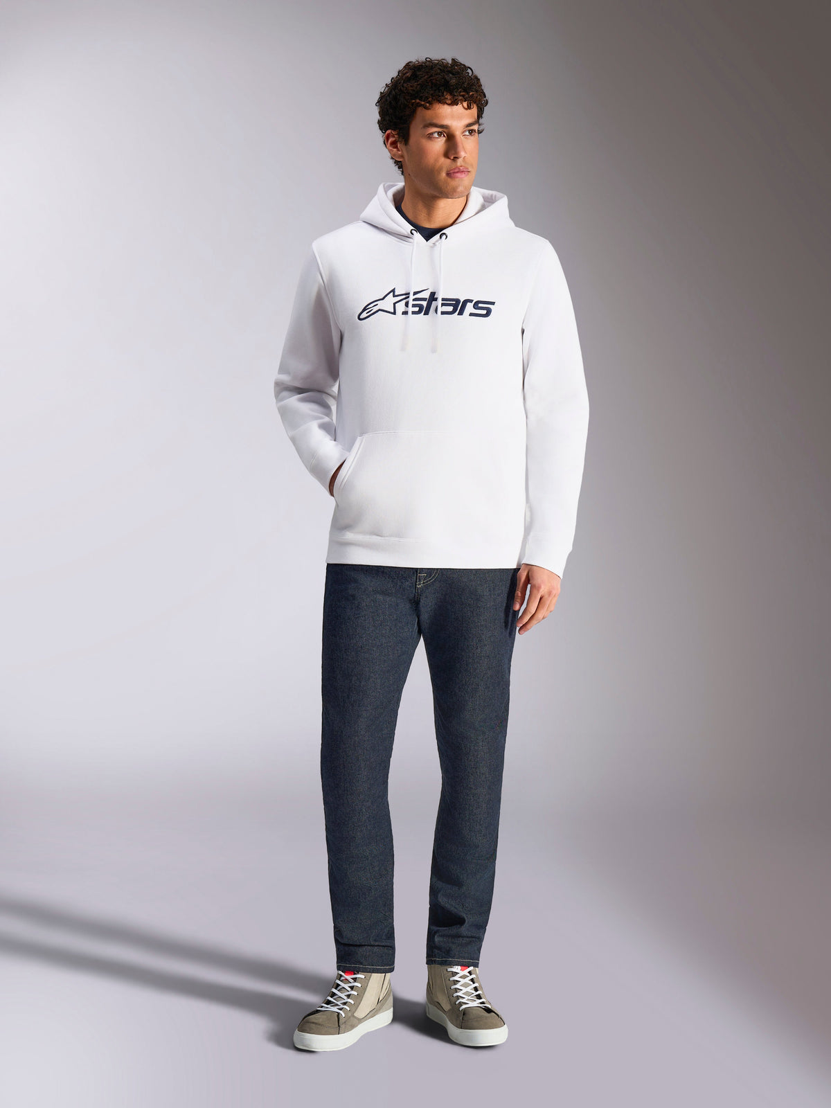 Sweat à capuche Alpinestars Blaze V3, sweat à capuche blanc de style streetwear décontracté avec logo Alpinestars bleu marine sur la poitrine, porté par un mannequin avec un jean bleu foncé et des baskets beiges, doté d'une capuche à cordon de serrage et d'une poche kangourou à l'avant