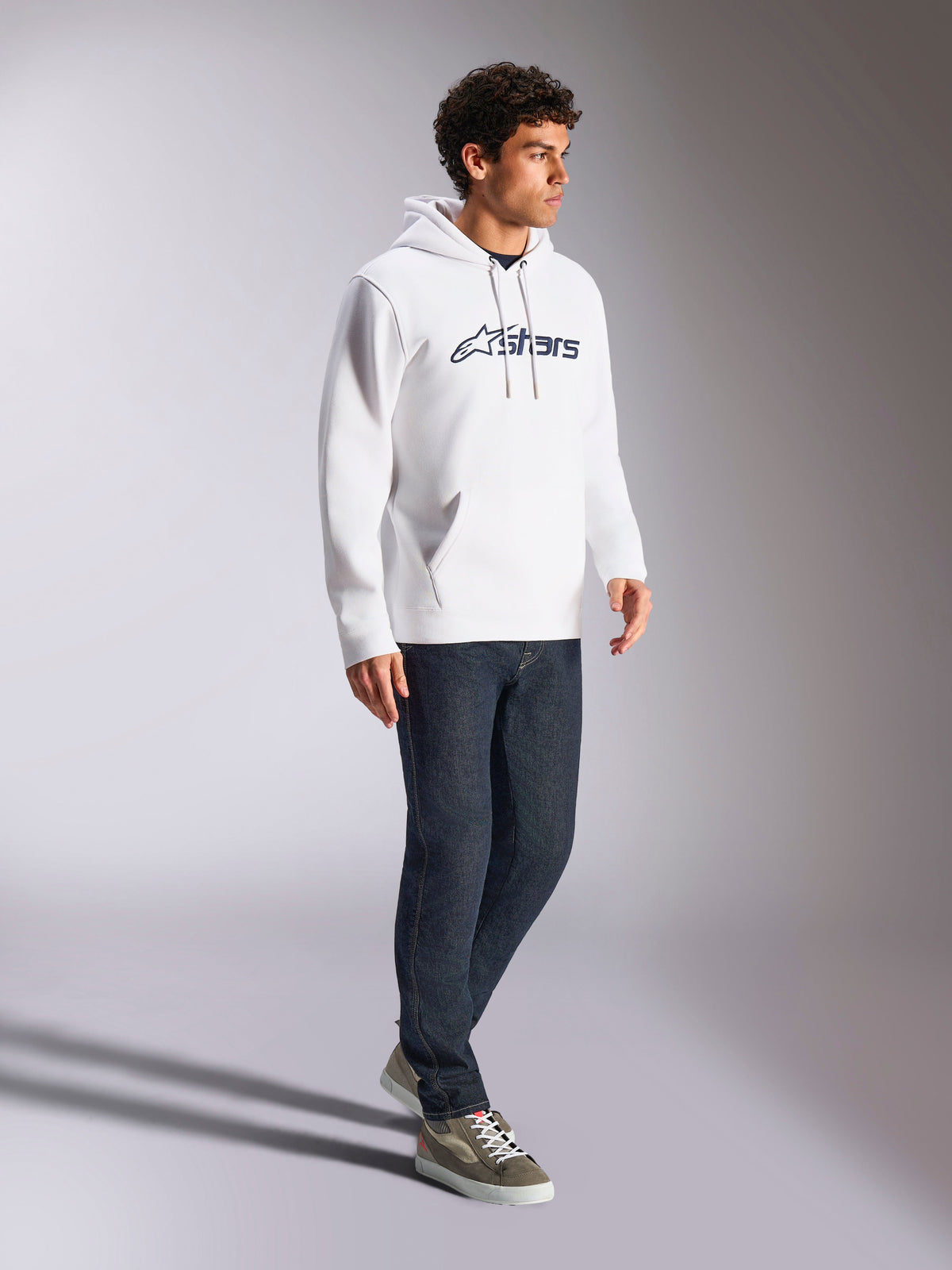 Sweat à capuche Alpinestars Blaze V3, blanc et bleu marine, tenue streetwear décontractée portée par un mannequin avec un jean bleu foncé et des baskets, comprend une capuche à cordon de serrage et un logo imprimé audacieux à l'avant, design moderne inspiré des sports mécaniques