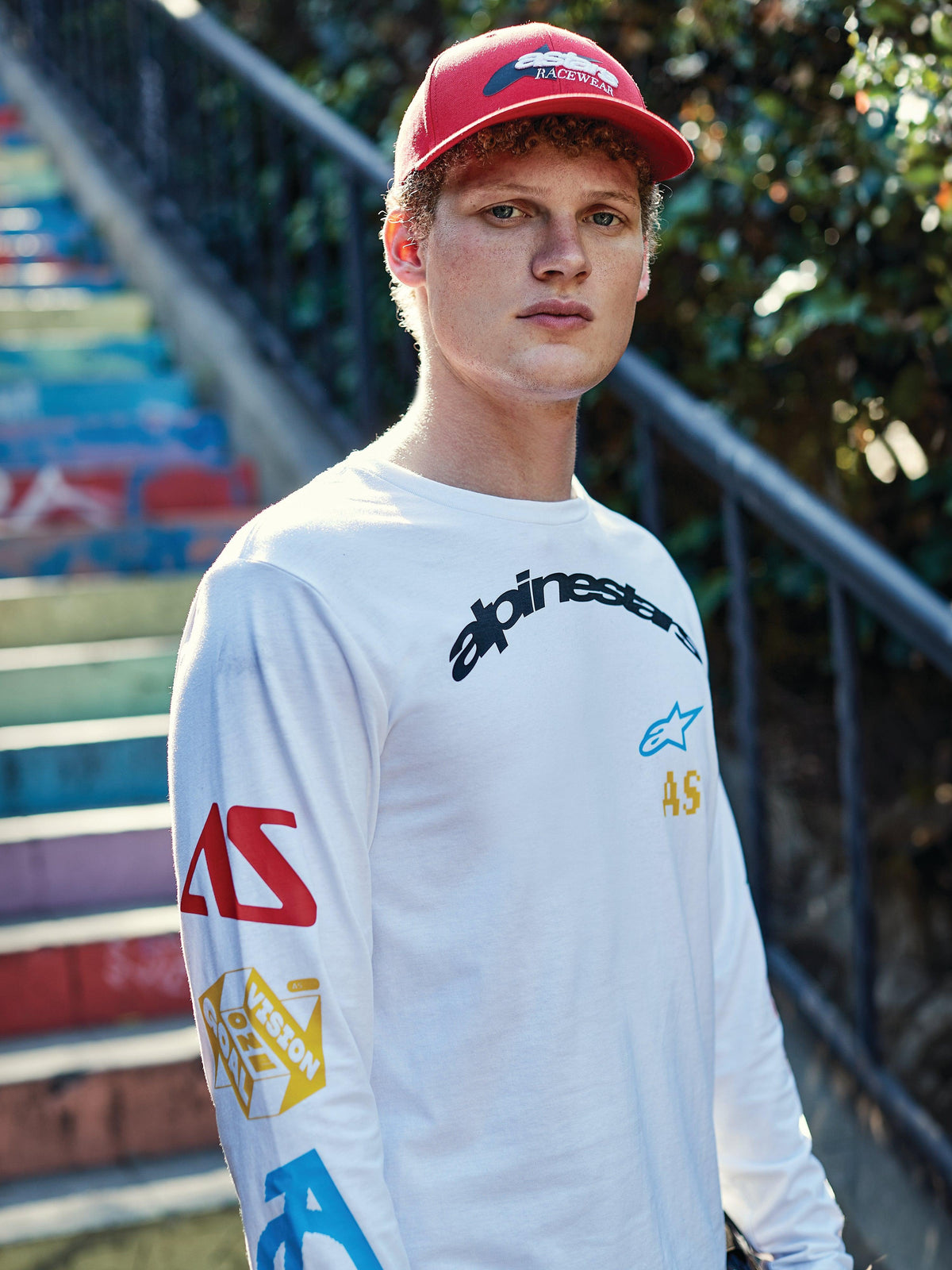 Alpinestars Profile Mütze, Rot, getragen von einem jungen Mann, strukturierte Krone mit gebogenem Schirm und besticktem Logo, kombiniert mit einem weißen Langarm-T-Shirt mit Grafik, lässige Motorsport-Streetwear