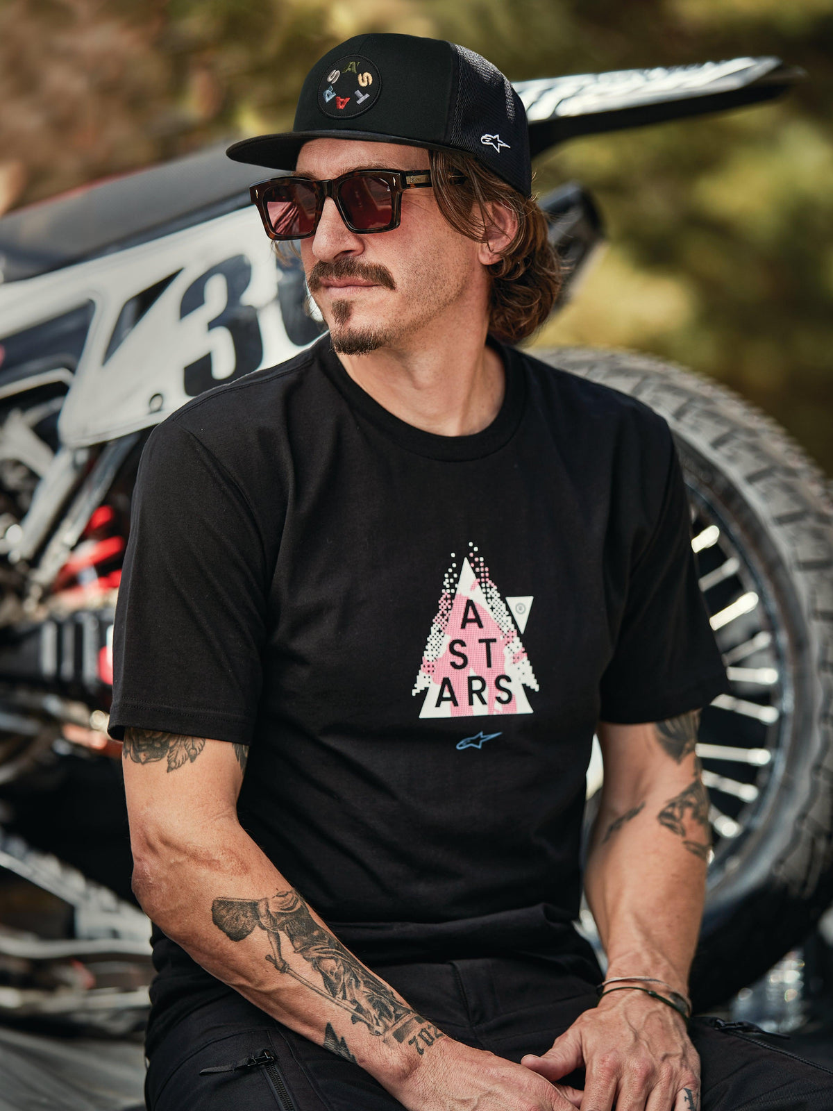 Alpinestars Gambit Trucker Mütze in Schwarz mit kreisförmigem Logo-Patch, getragen mit Alpinestars Soloist SS T-Shirt in Schwarz, Lifestyle-Aufnahme eines Mannes mit Sonnenbrille und Tätowierungen vor einem Motocross-Motorrad.