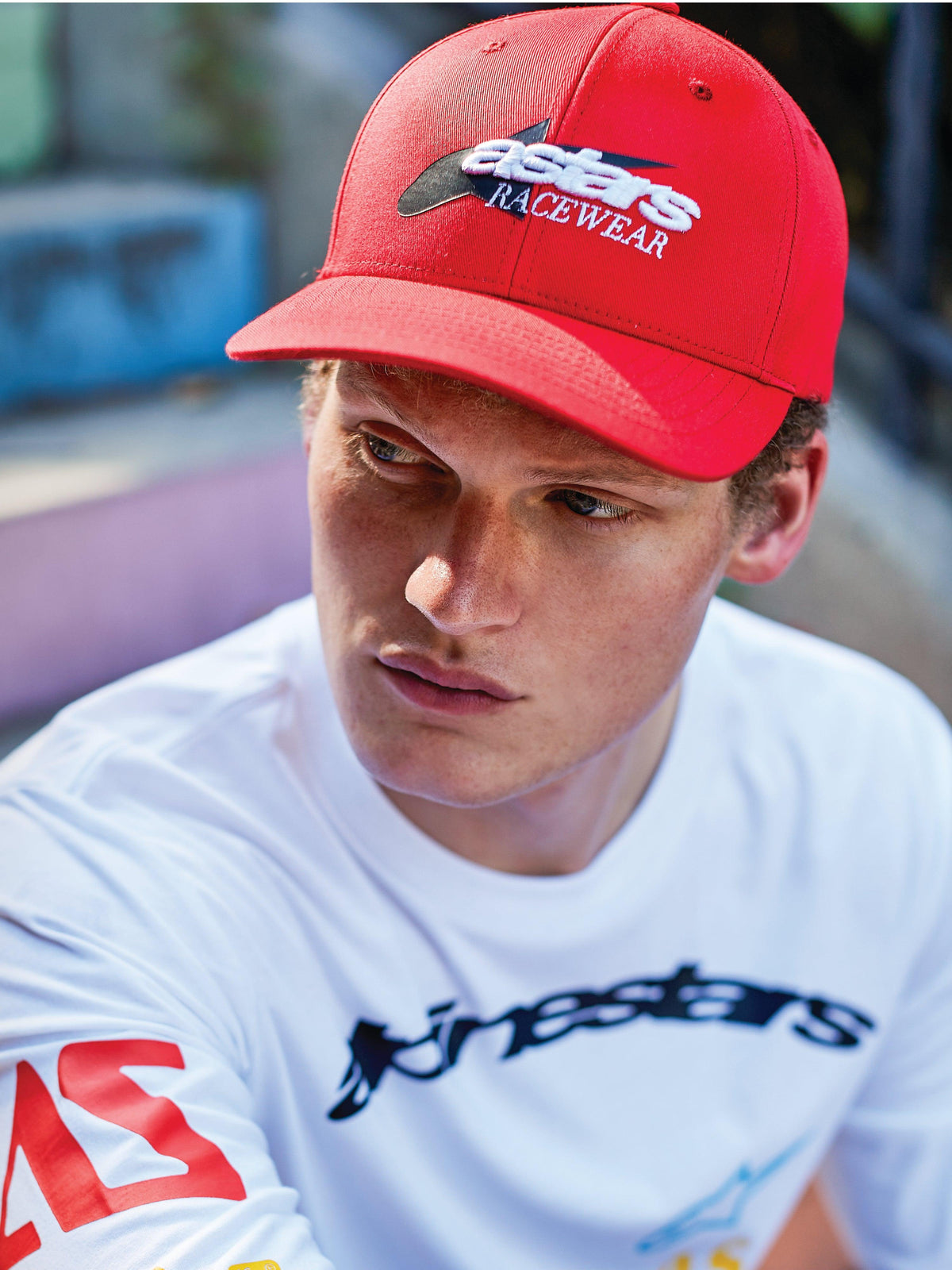Mann mit Alpinestars Profile Mütze, rote Snapback-Kappe mit gebogenem Schirm, weiß-schwarze „astars RACEWEAR“ Frontstickerei, kombiniert mit einem weißen Alpinestars Langarm-Grafik-T-Shirt, lässiger Motorsport-Lifestyle-Look