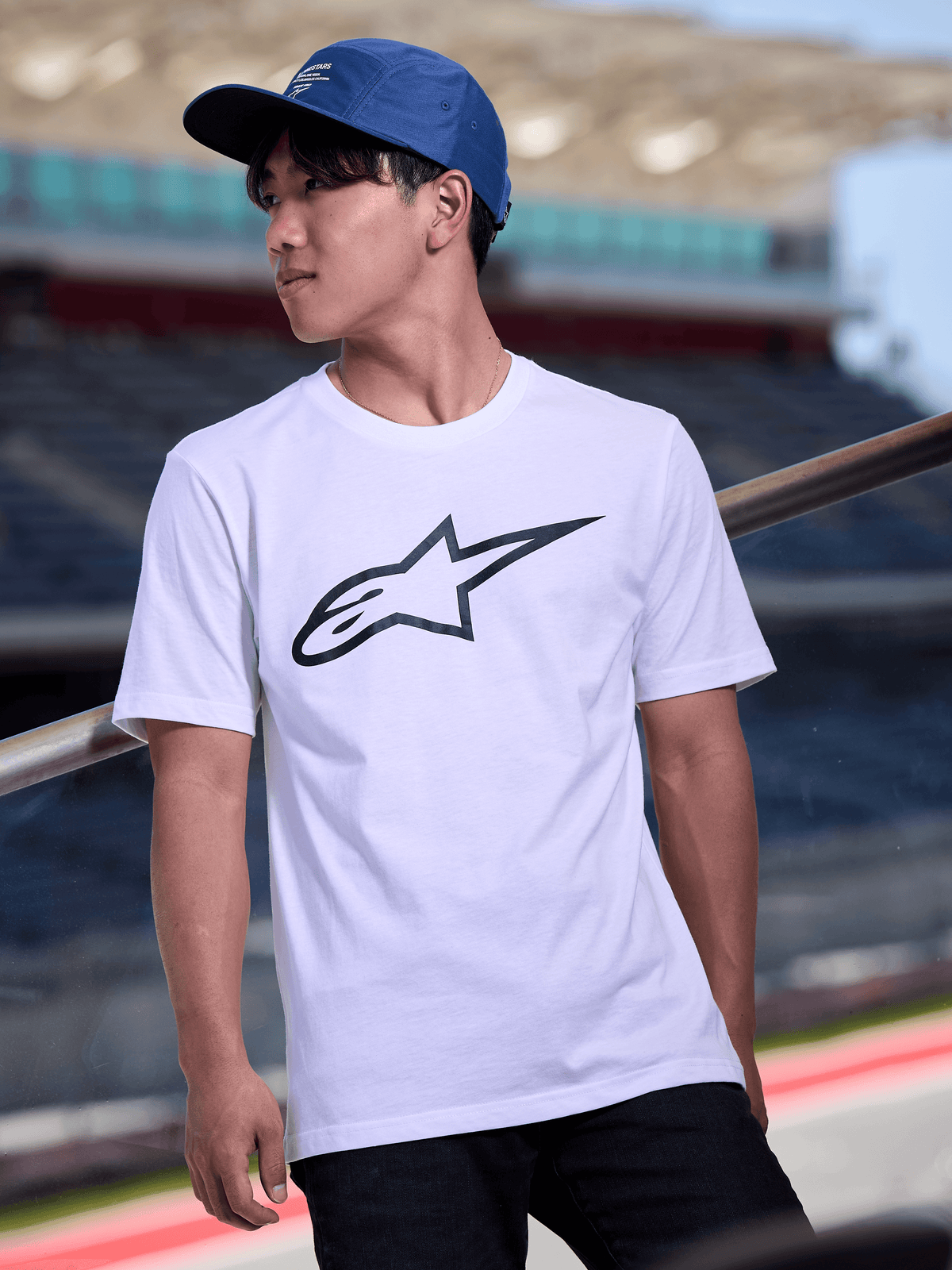 Mann trägt Alpinestars Peasy Mütze in Blau, 5-Panel-Jockey-Kappe mit flachem Schirm und weißem Grafik-Logo-Druck, kombiniert mit einem weißen Alpinestars Logo-T-Shirt, lässige, vom Motorsport inspirierte Streetwear