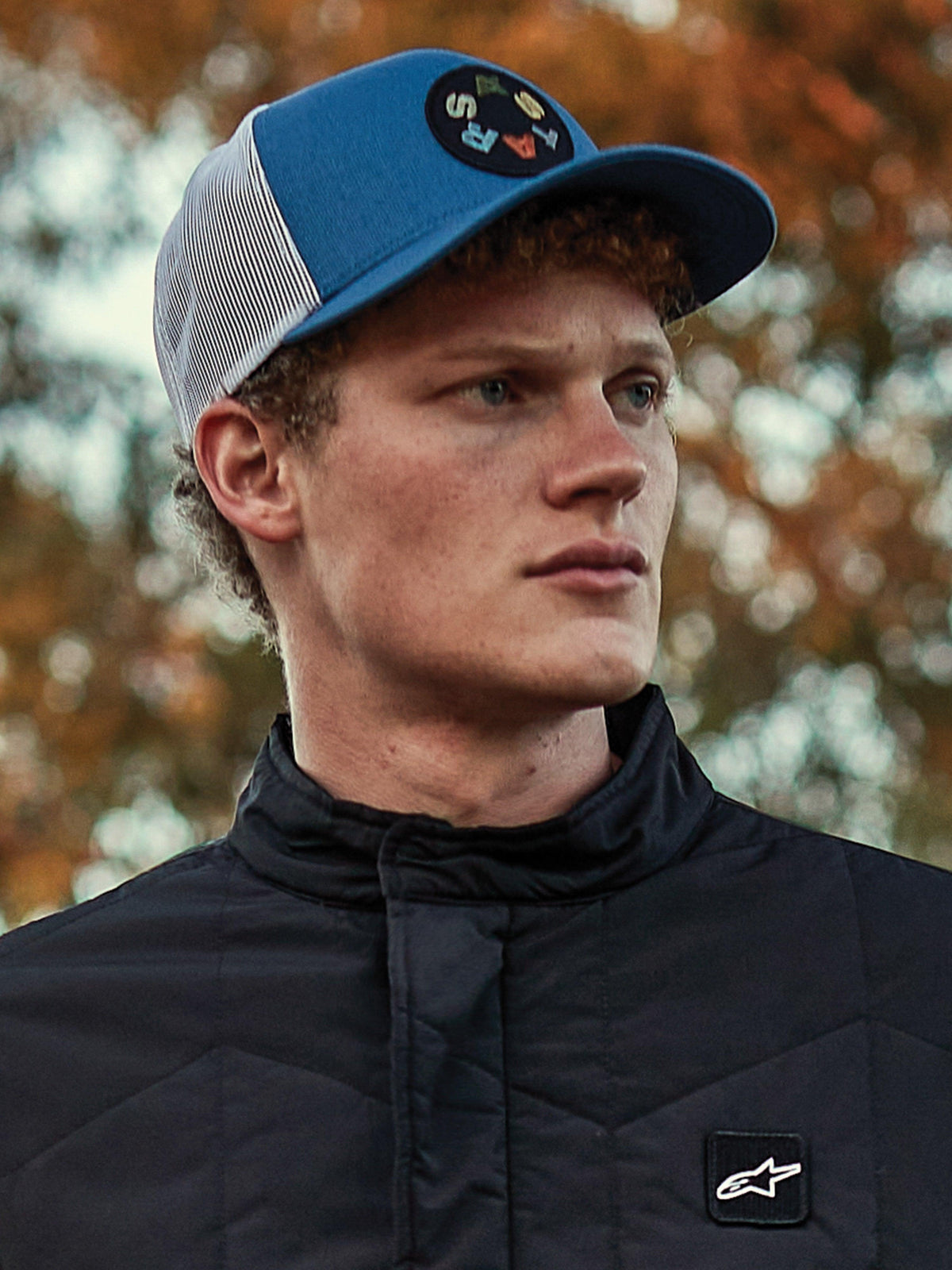 Mann mit Alpinestars Gambit Trucker Hat, blau-weiße Cap mit Mesh-Rückseite und kreisförmigem Astars-Logo-Patch, kombiniert mit einer schwarzen gesteppten Alpinestars Jacke, lässiges Motorsport-Lifestyle-Porträt vor herbstlichem Hintergrund