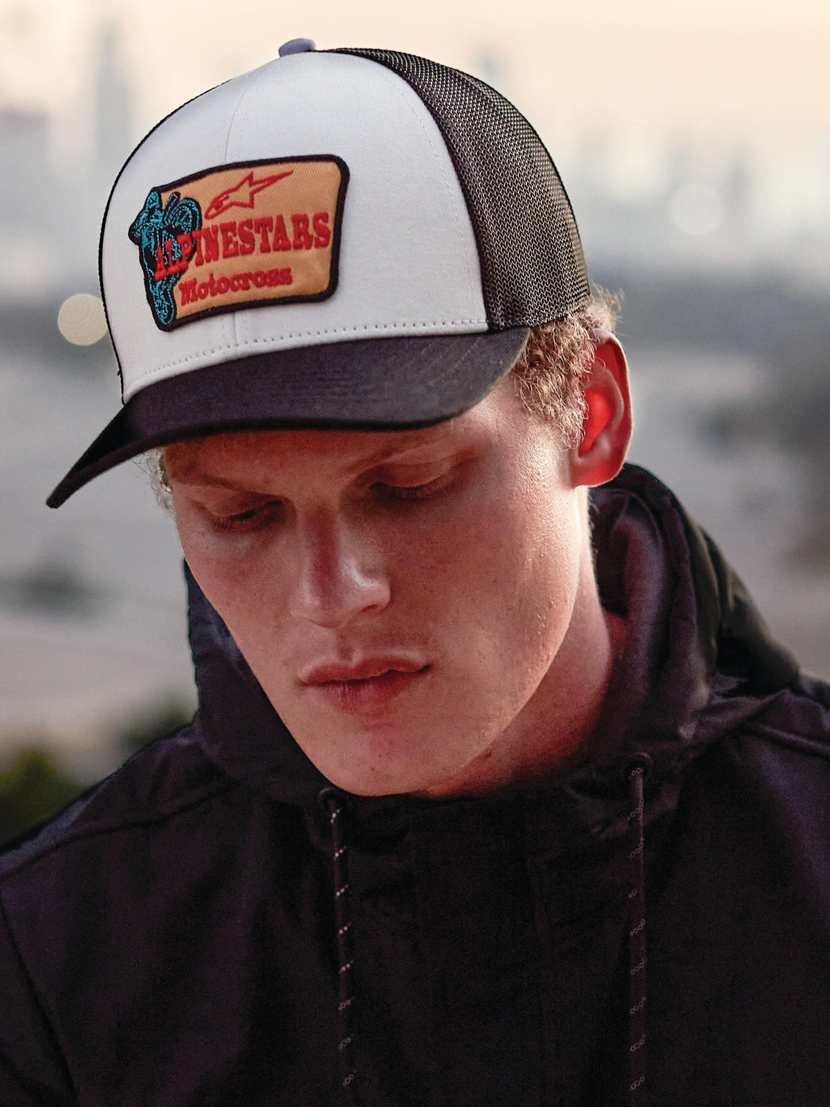 Casquette Trucker Alpinestars Hardware en Military White, portée par un homme en sweat à capuche noir, dotée d'un panneau avant blanc avec visière courbée noire et arrière en maille noire, écusson brodé beige avec marquage Alpinestars Motocross rouge, style lifestyle motorsport streetwear décontracté