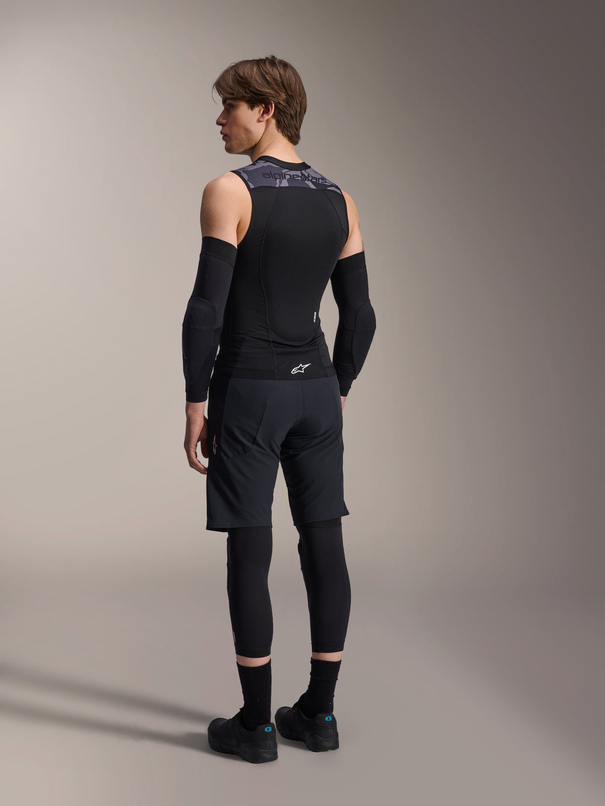 Mannelijk model van de achterkant in Alpinestars A-Motion Plasma Protection Vest, Black Camo, mouwloos mesh-ontwerp met geïntegreerde rugbeschermer, grijs camouflagepatroon op de schouders, gedragen met zwarte elleboogbeschermers en mountainbikeshorts, professionele wieleruitrusting