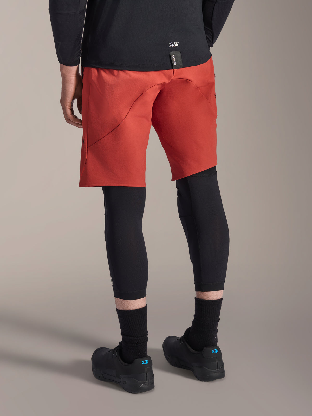 Alpinestars A-Motion Plasma Knee Sleeve, zwarte MTB kniebeschermer, gedragen met een rode mountainbike short en zwarte fietsschoenen, achteraanzicht toont het onopvallende slip-on ontwerp voor off-road fietsen
