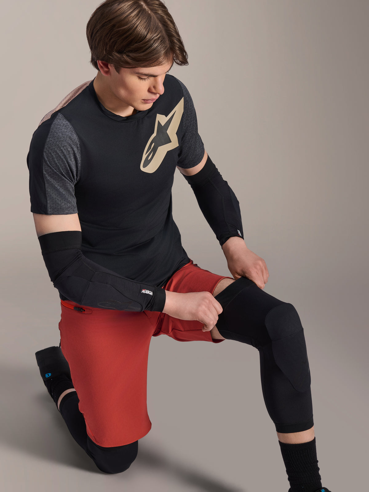 Alpinestars A-Motion Plasma Knee Sleeve, MTB-kniebeschermer, zwart, gedragen door een rijder in een knielende positie, gecombineerd met een zwart en grijs T-shirt met een goudkleurig logo, rode short en zwarte elleboogbeschermers, professionele mountainbike beschermingsuitrusting