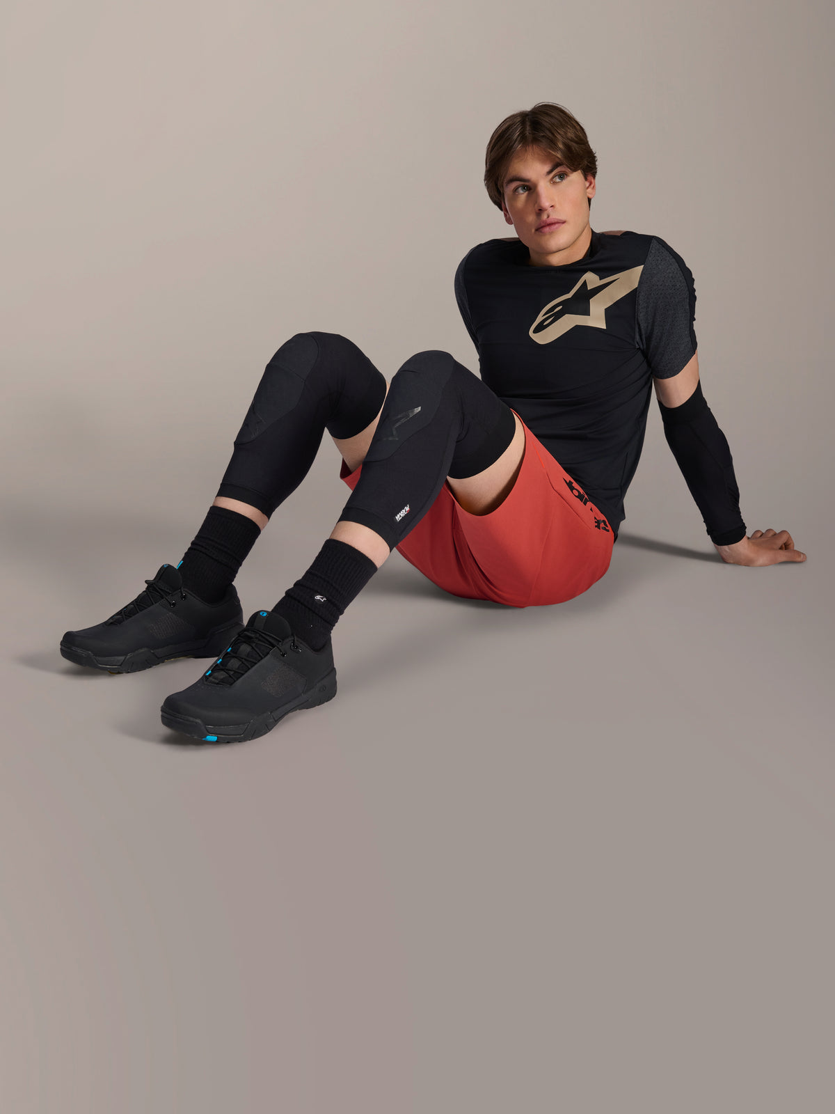 Alpinestars A-Motion Plasma Knee Sleeve, zwarte mountainbike kniebeschermers, gedragen door een mannelijk model zittend in een complete fietsuitrusting met een rode short en een zwart technisch T-shirt, toont het low-profile beschermende ontwerp.