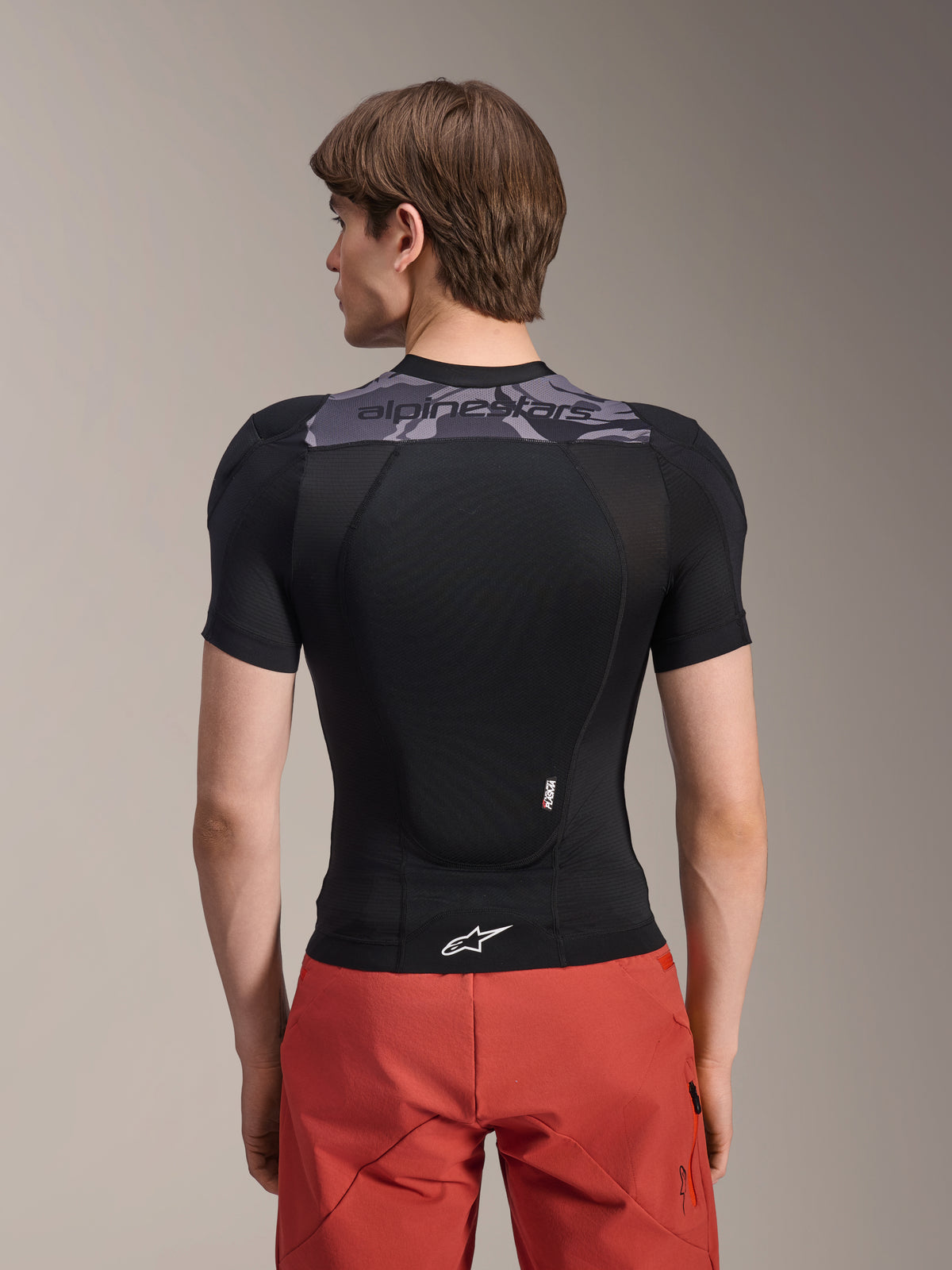Alpinestars A-Motion Plasma Protection Jacket - Korte Mouwen, zwart met grijze camo-accenten op de bovenrug, achteraanzicht met geïntegreerde rugprotector en wit Astars-logo op de zoom, gedragen met rode short, technische mountainbike-beschermingsuitrusting