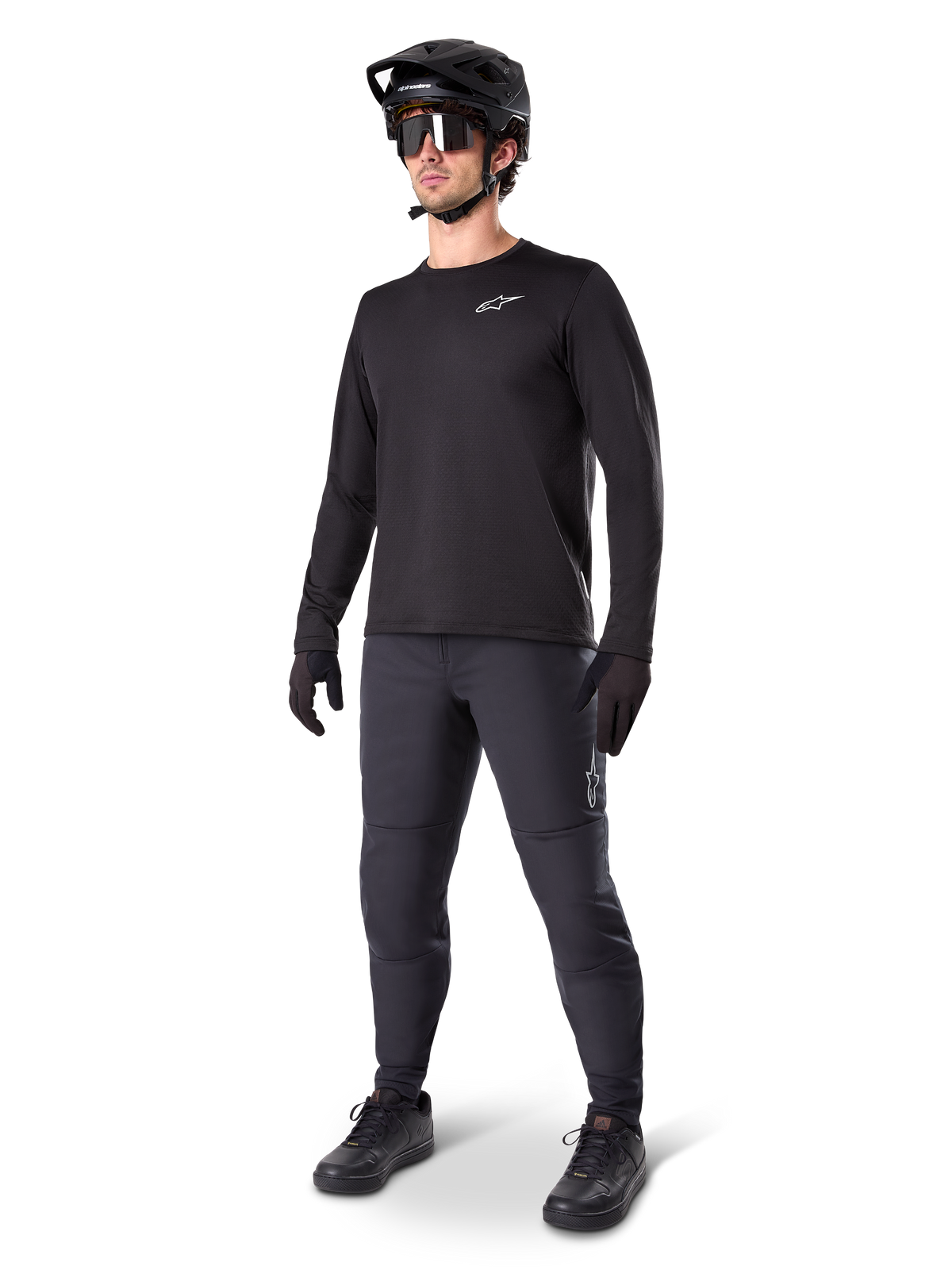 Cycliste de VTT portant le pantalon thermique Alpinestars A-Dura noir, avec un maillot à manches longues noir, un casque noir et des lunettes de soleil, logo Alpinestars argenté sur la cuisse gauche, conçu pour le trail par temps froid.