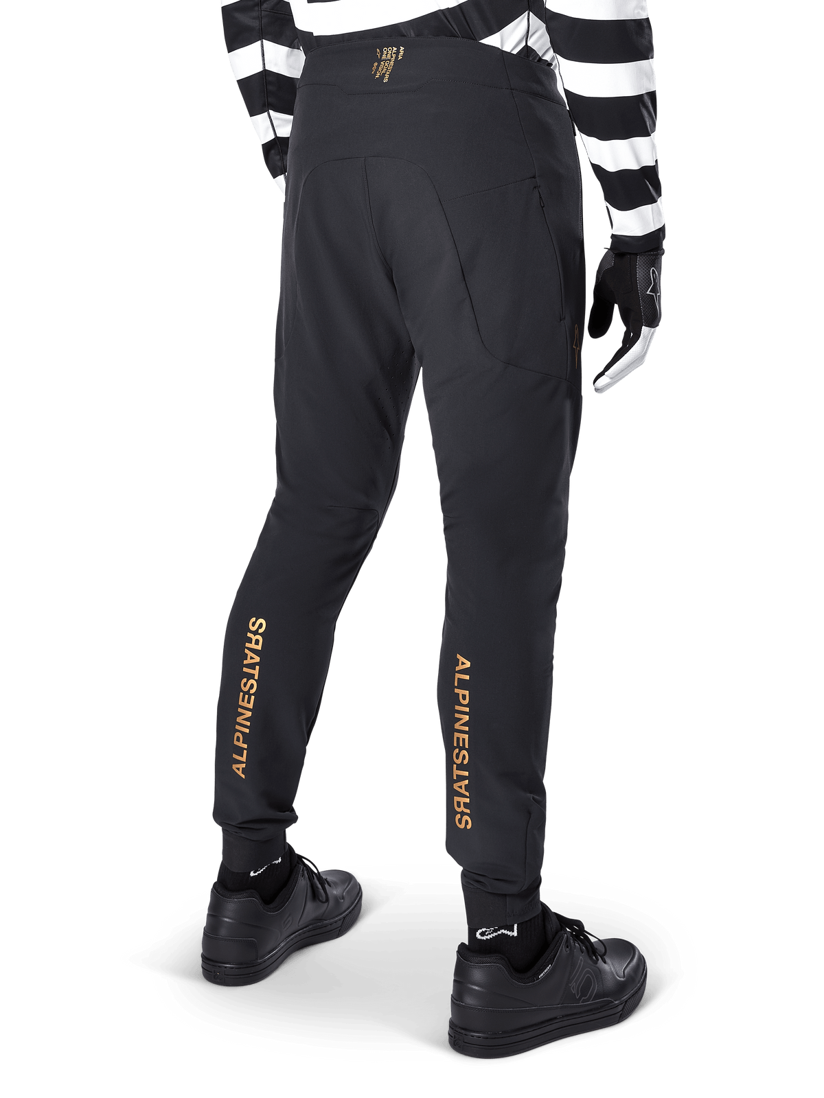 Alpinestars A-Aria Elite Hose, schwarze MTB-Hose, Rückansicht, mit vertikalem goldenem Alpinestars-Branding auf der Rückseite der Beine, getragen mit einem schwarz-weiß gestreiften Trikot und schwarzen Schuhen, Performance-Radbekleidung für Mountainbiking