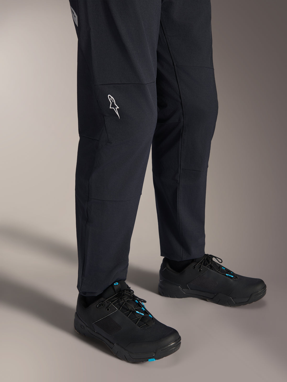 Alpinestars A-Dura Elite Hose, schwarze Mountainbike-Hose, Detailansicht der unteren Beine mit weißem Alpinestars-Logo am Knie, getragen mit schwarz-blauen Fahrradschuhen, strapazierfähige Konstruktion für MTB