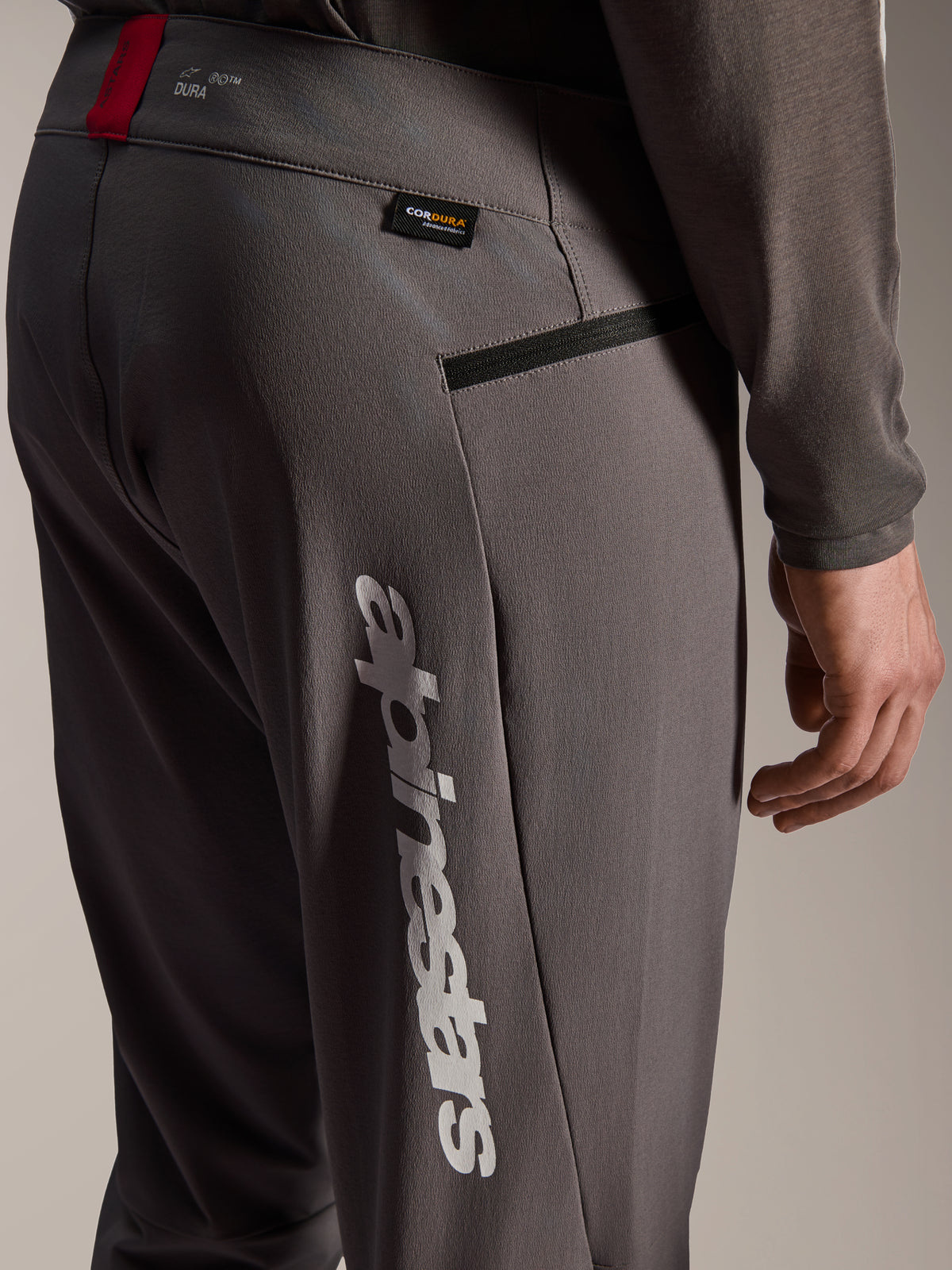 Alpinestars A-Dura Elite Pants, donkergrijze MTB-broek, close-up van de heup en zijkant van het bovenbeen, met groot wit verticaal Alpinestars-logo, Cordura-label, zak met ritssluiting en rood accent op de tailleband