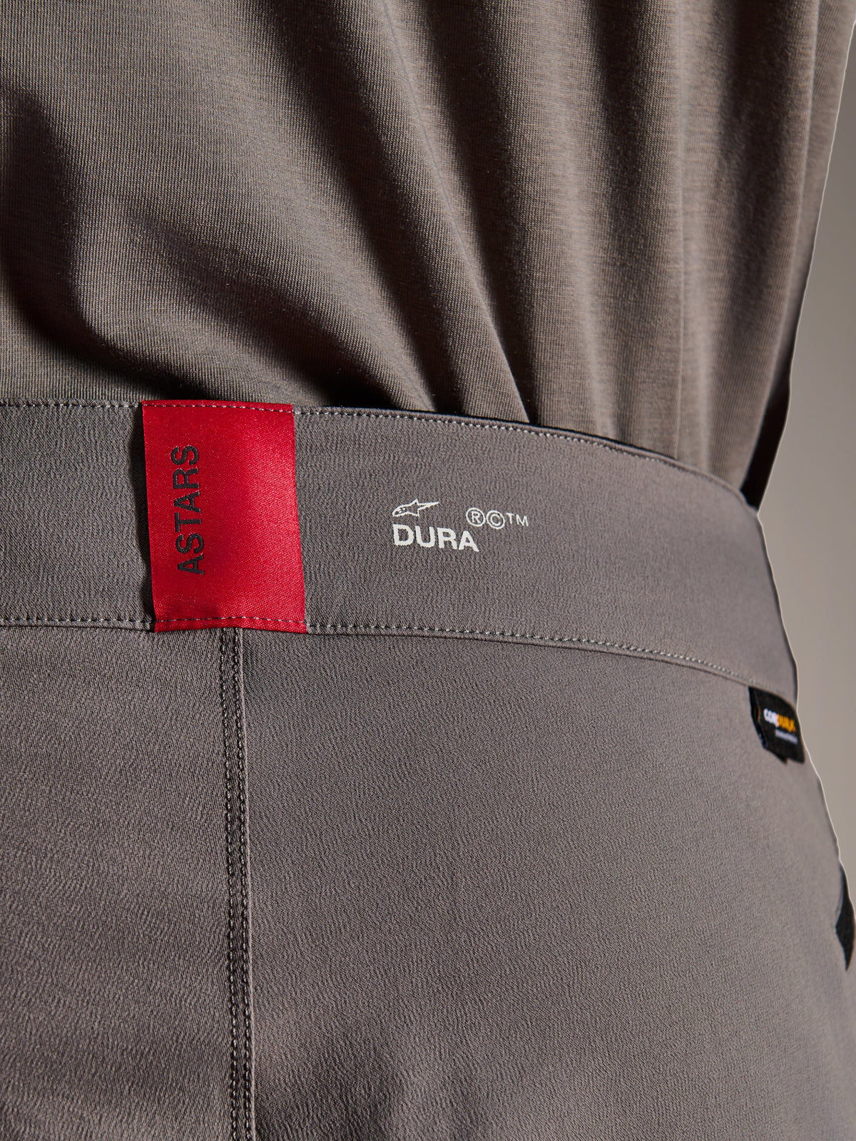 Alpinestars A-Dura Elite Pants, MTB-broek, donkergrijs, close-up van de tailleband met een rood ASTARS-label en wit DURA-logo, voorzien van getextureerde Cordura-stof en hoogwaardige brandingdetails.