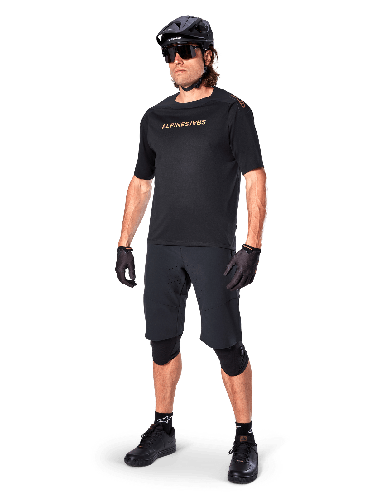 Fietser die Alpinestars A-Aria Elite shorts draagt, zwarte mountainbikebroek, gecombineerd met een bijpassende zwarte jersey met gouden branding, zwarte helm, zonnebril en kniebeschermers, complete performance-outfit voor off-road fietsen