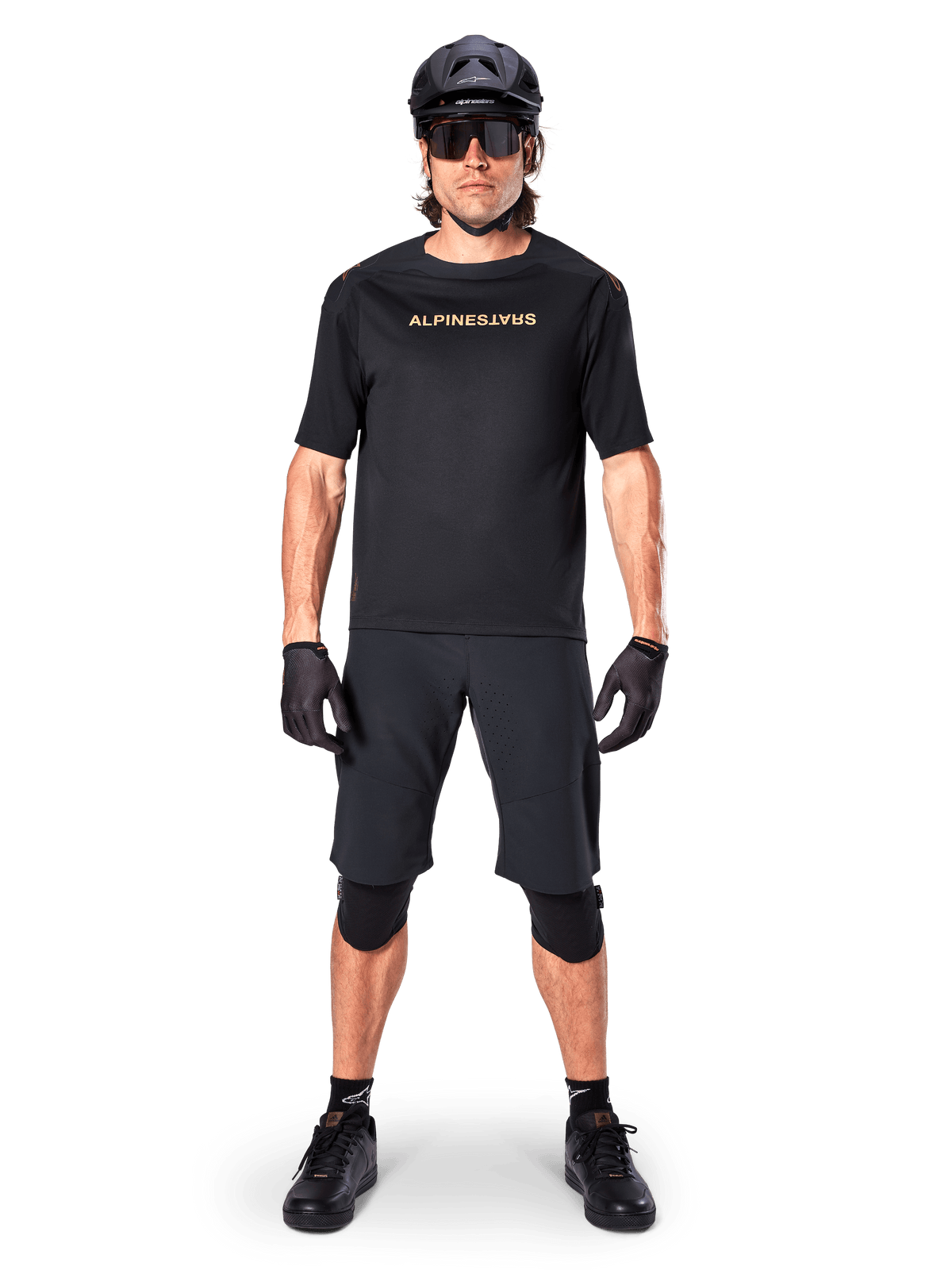 Mannelijke mountainbiker die zwarte Alpinestars A-Aria Elite Shorts draagt, gecombineerd met een bijpassend zwart Alpinestars fietsshirt met korte mouwen, zwarte helm, handschoenen en kniebeschermers, complete off-road wieleroutfit