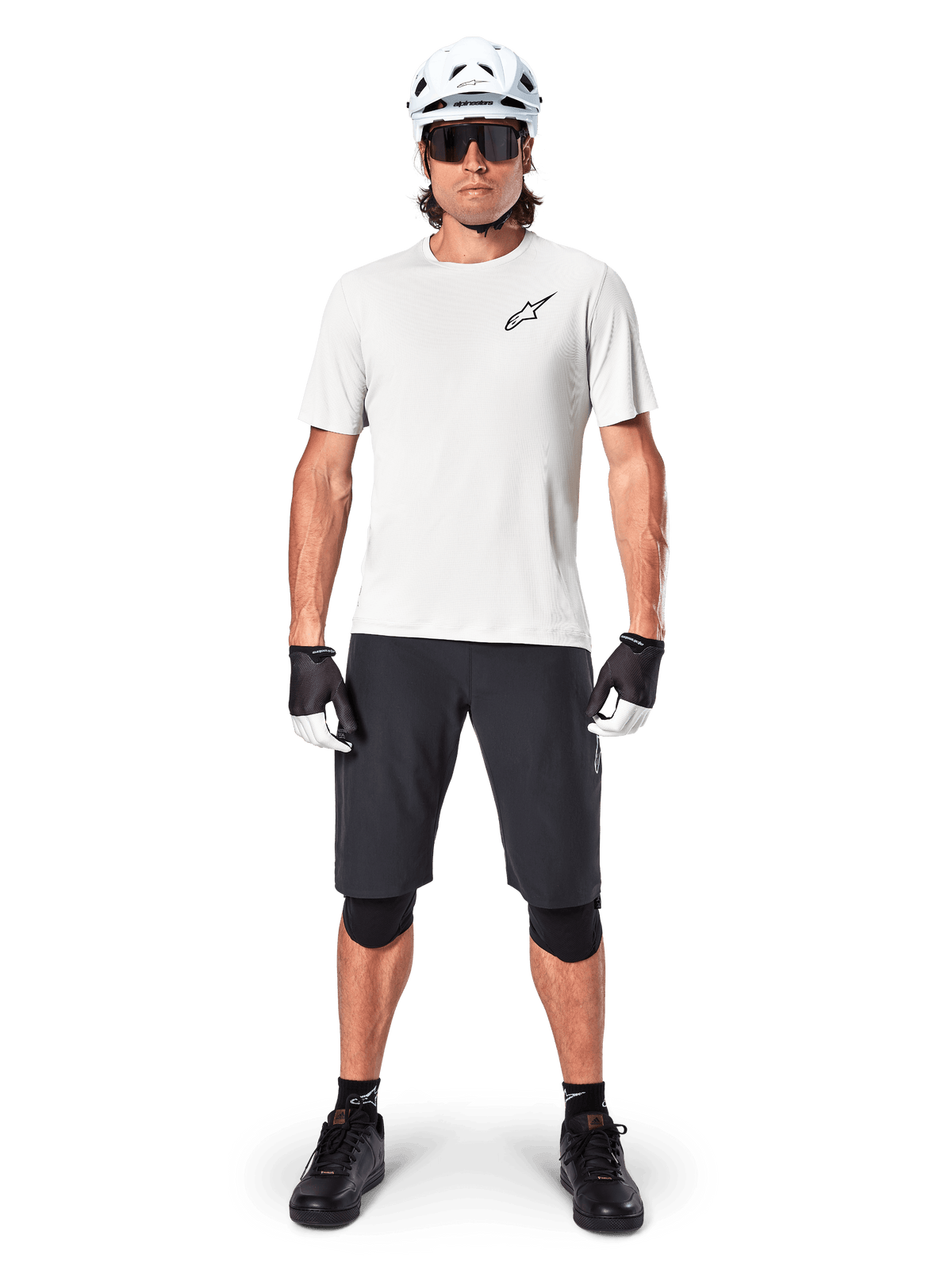 Mannelijke mountainbiker met Alpinestars A-Aria Shorts, zwart, gecombineerd met een wit performance fietsshirt, witte helm en zwarte kniebeschermers, professionele off-road fietsuitrusting in een volledig vooraanzicht