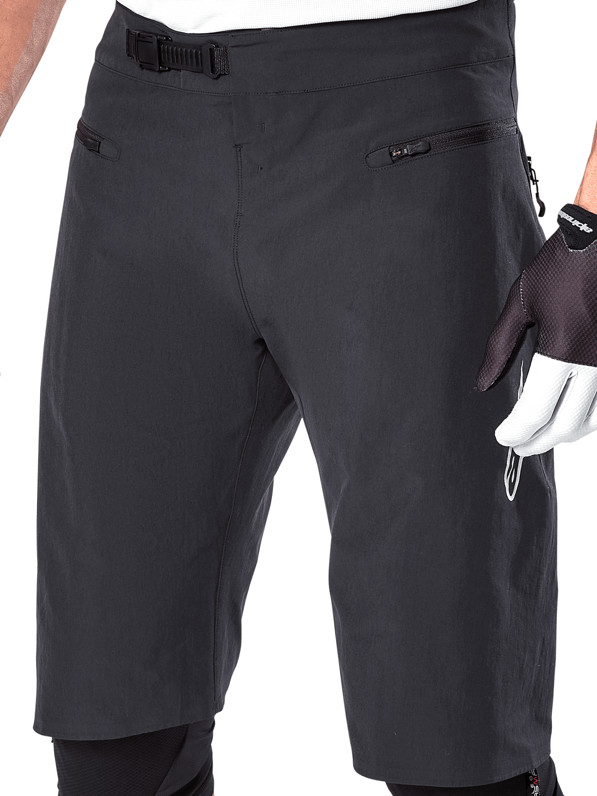 Alpinestars A-Aria Shorts, mountainbike shorts, zwart, close-up van de voorkant van de taille en dijen met het ratelsluitingssysteem en zakken met ritssluiting, gedragen met zwart-witte fietshandschoenen, performance trailuitrusting.