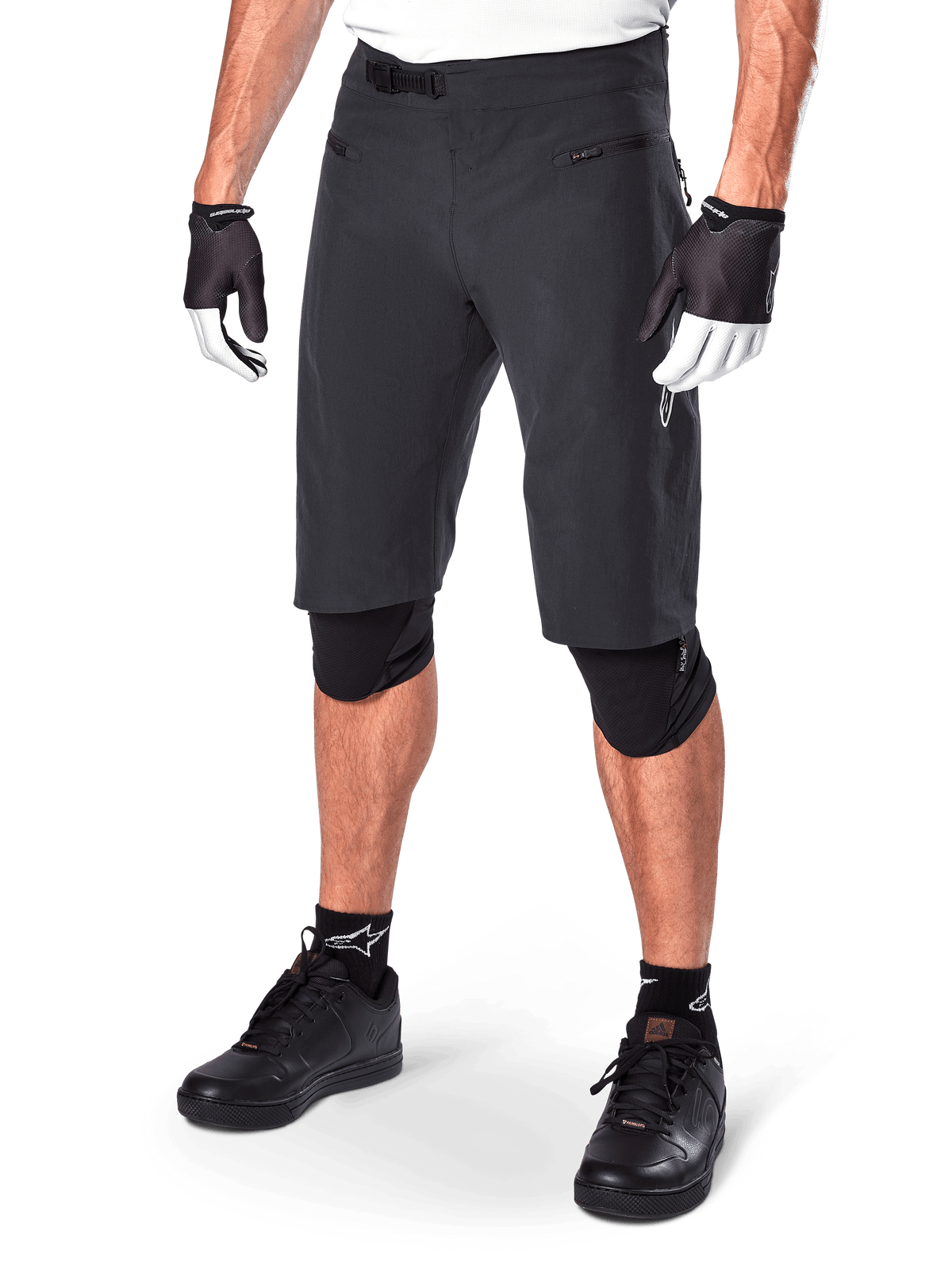 Alpinestars A-Aria Shorts, mountainbike shorts, zwart, knielange pasvorm met ritszakken aan de voorzijde en verstelbare ratelsluiting in de taille, gedragen met zwarte kniebeschermers, sokken en fietshandschoenen