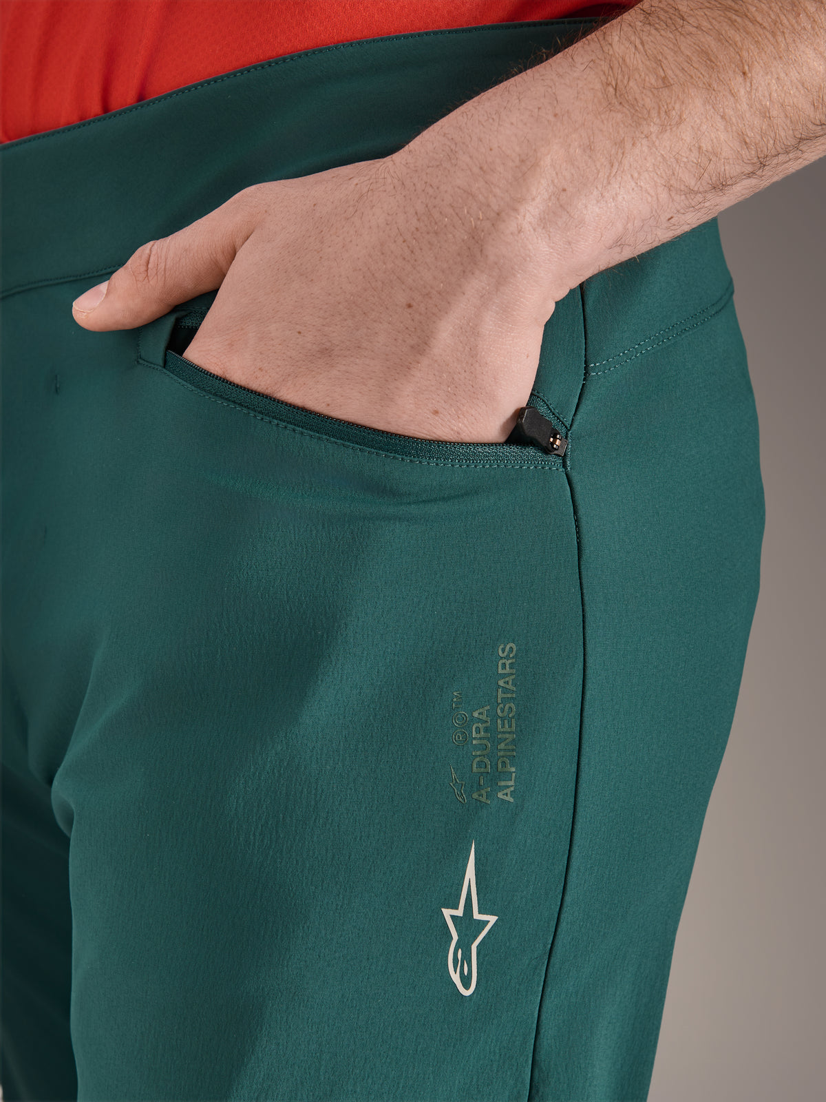 Alpinestars A-Dura Pants in Dark Pine groen, close-up van zak met ritssluiting en hand, voorzien van wit Alpinestars-logo en tekstbranding op het bovenbeen, duurzame MTB-fietsbroek