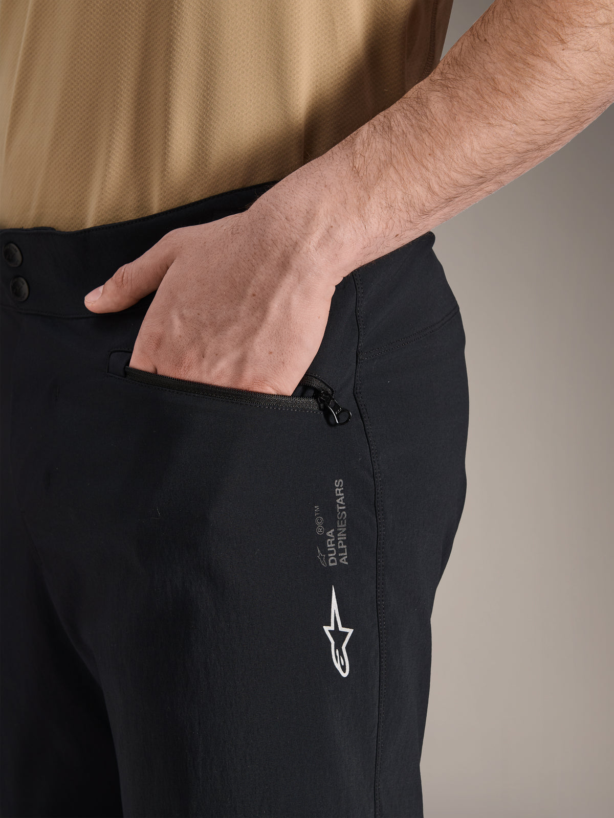 Short Alpinestars A-Dura, short de VTT noir, gros plan sur la poche latérale zippée avec une main à l'intérieur, arborant le logo Alpinestars blanc et le marquage A-Dura, textile durable pour le cyclisme tout-terrain