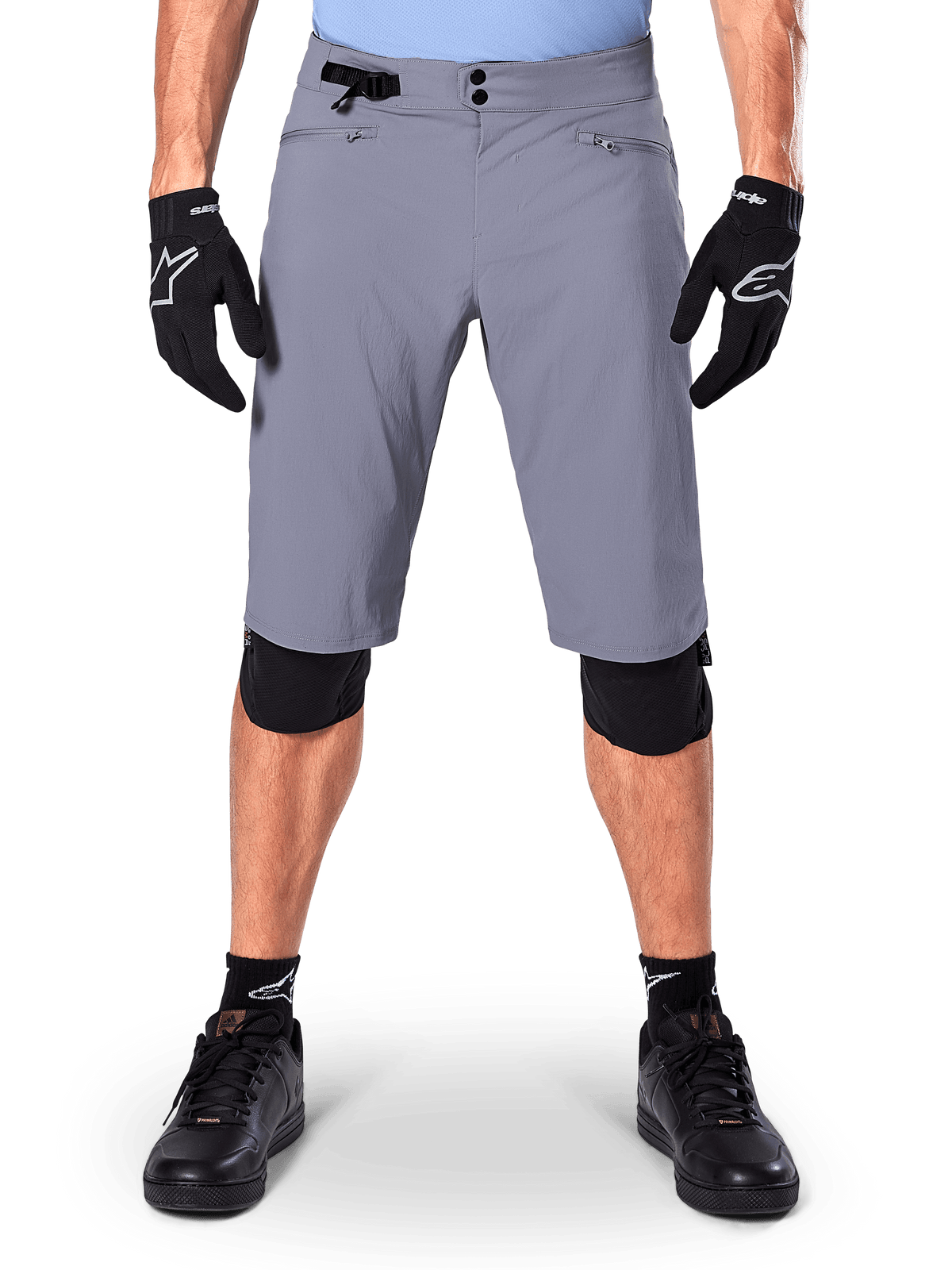 Alpinestars A-Dura shorts, mountainbikeshorts, donkergrijs, gedragen met zwarte Alpinestars-handschoenen en kniebeschermers, voorzien van een dubbele drukknoopsluiting en ritsbare, met mesh gevoerde zakken, duurzame 2-way stretchstof voor off-road fietsen